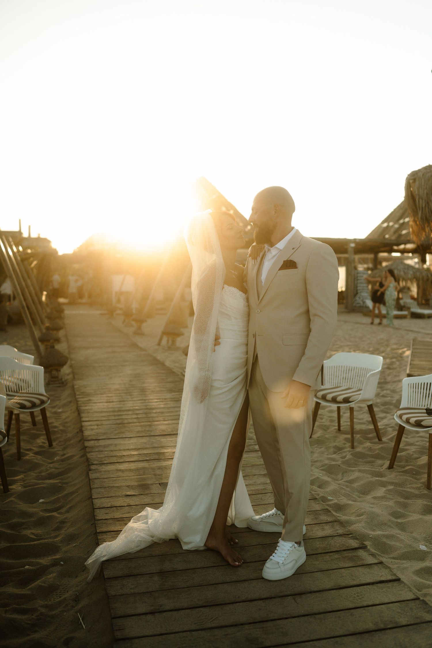Silhouettes of a couple dancing on a wooden deck by the beach during golden hour with chairs scattered around.