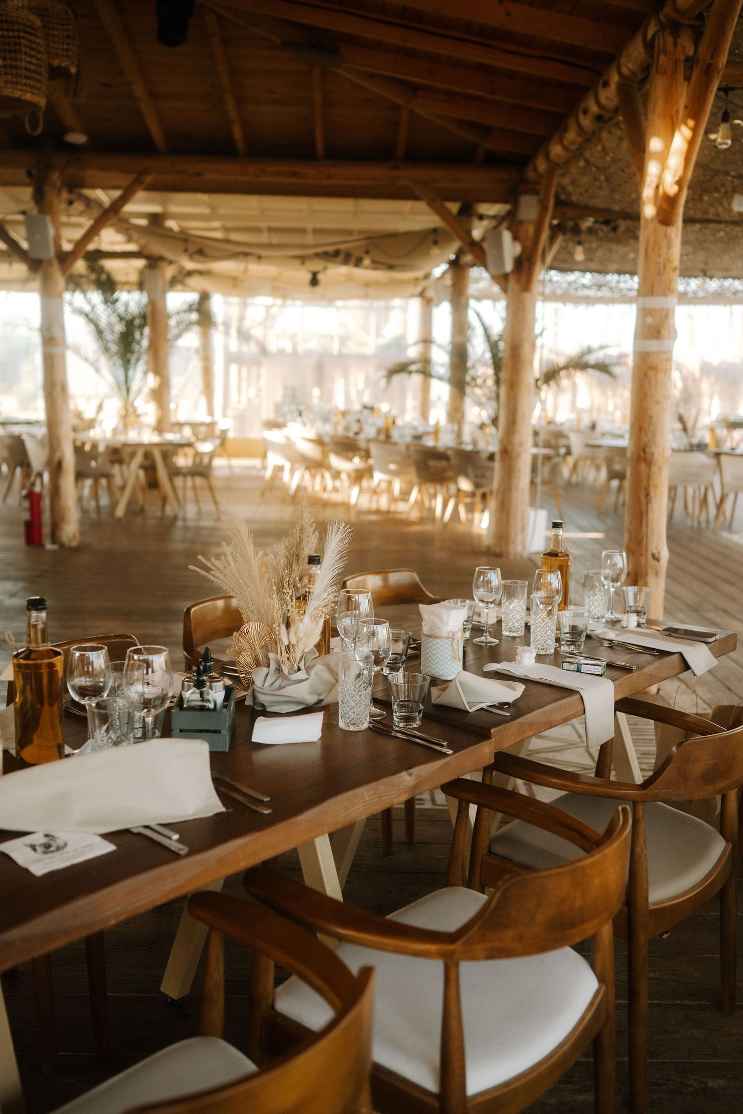 Rustic wedding reception venue with wooden tables and chairs decorated with dried wheat centerpieces in warm lighting.