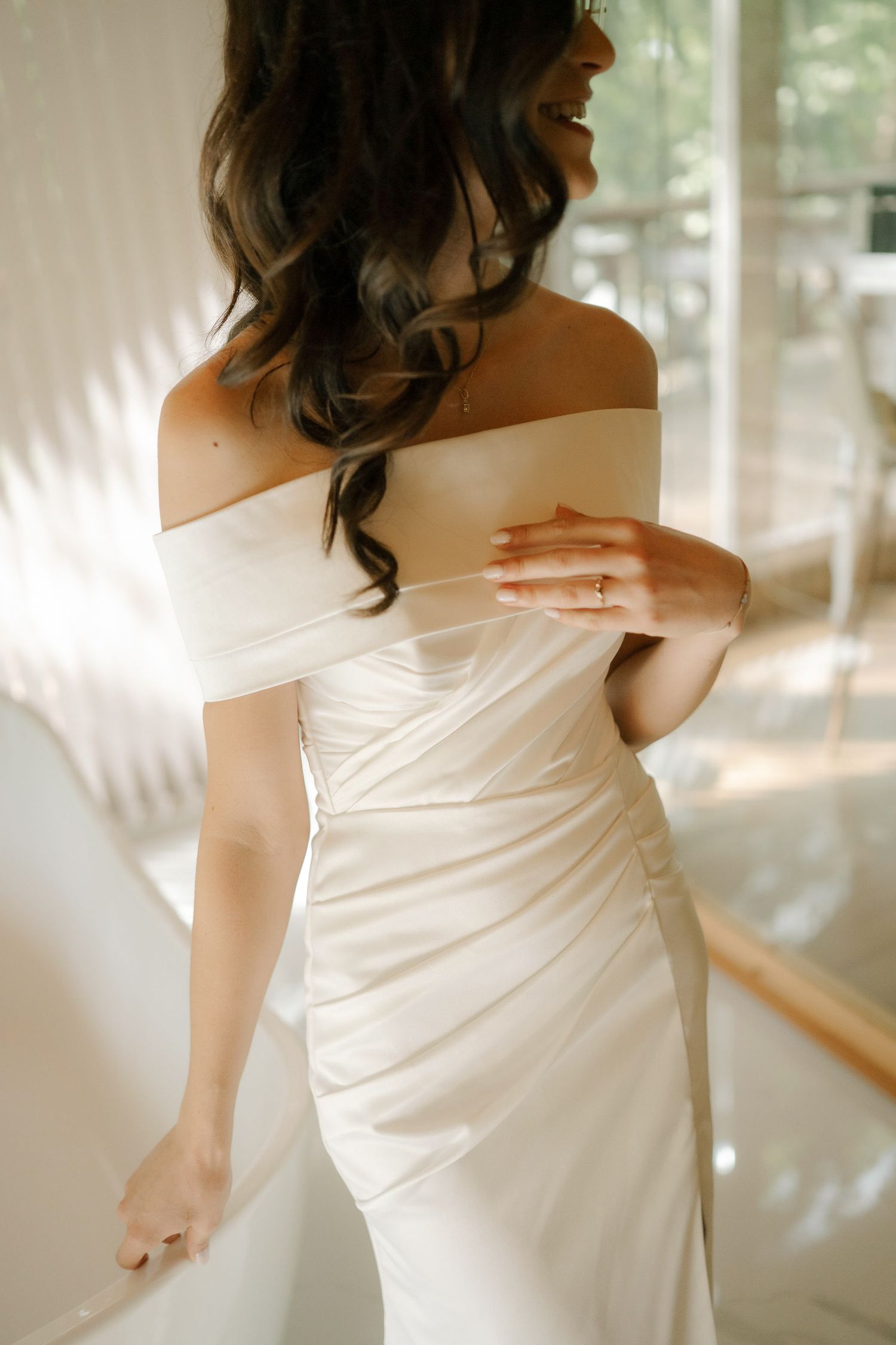 A bride in a white off-shoulder wedding dress is photographed from behind showing elegant details and flowing fabric.