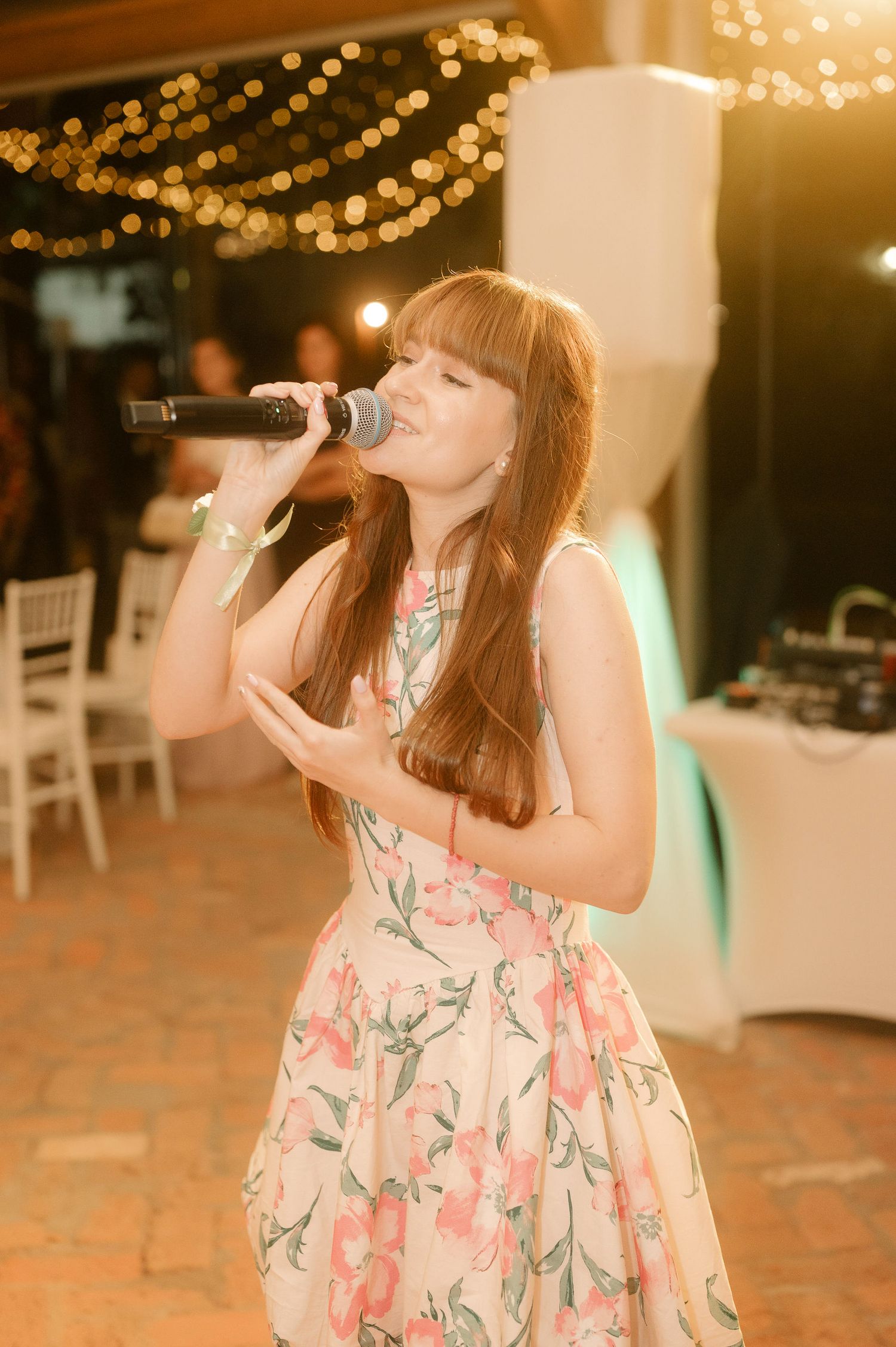 A person in a floral dress sips from a glass in a warmly lit venue with string lights overhead.