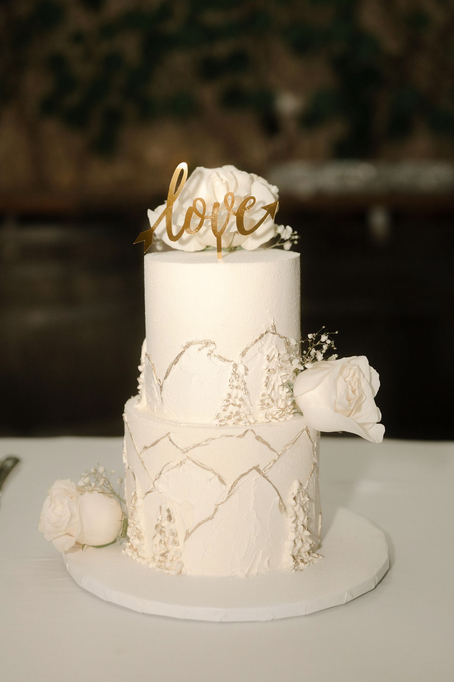 A white wedding cake with delicate lace details and gold accents displayed from multiple angles on a light surface.