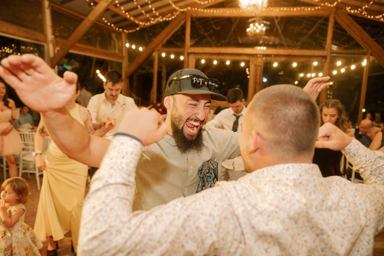 Two people dancing and celebrating at a rustic indoor wedding reception with string lights and wooden beams overhead.