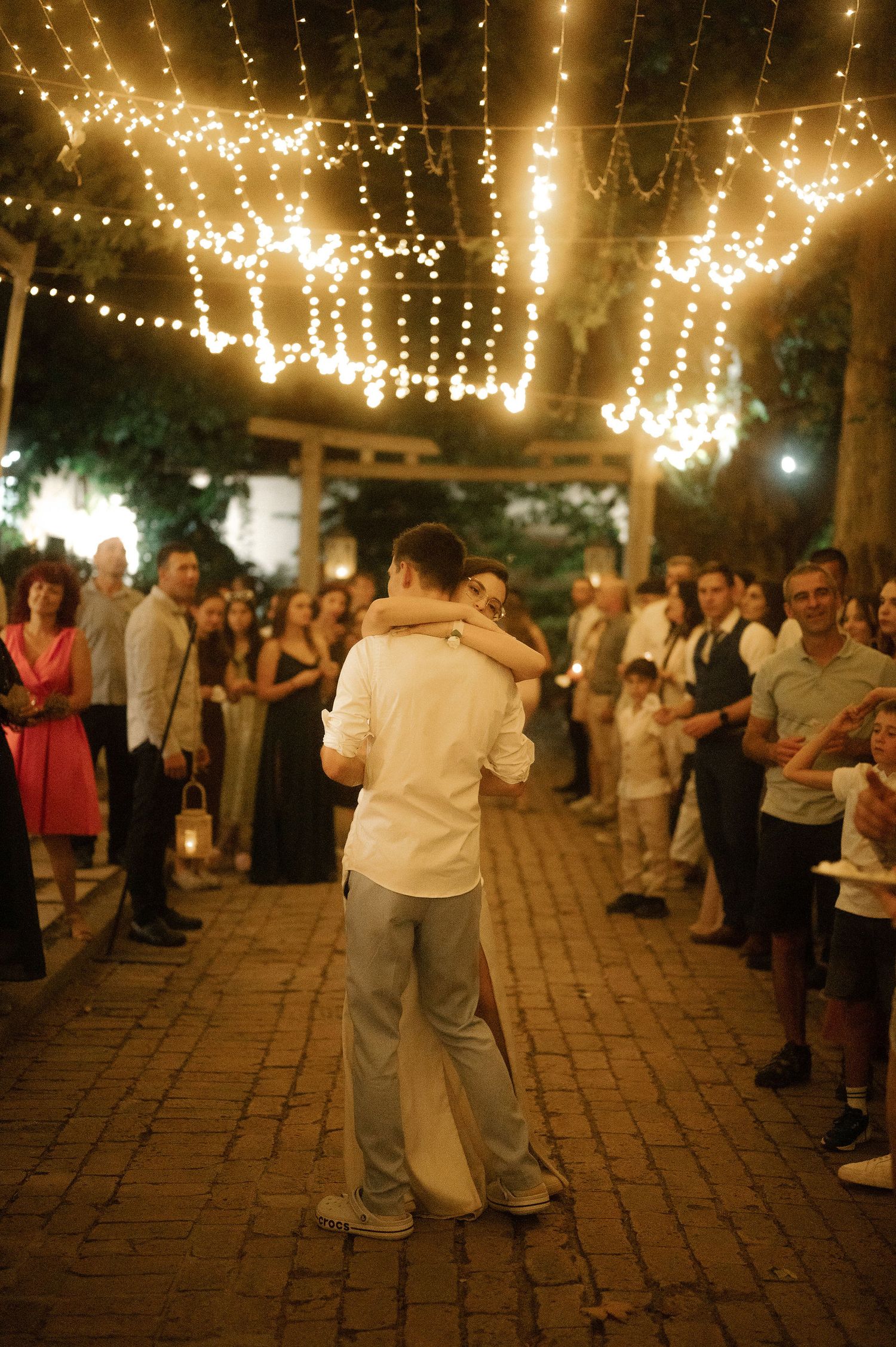 Couples dancing at an outdoor evening reception under romantic string lights on a brick patio.