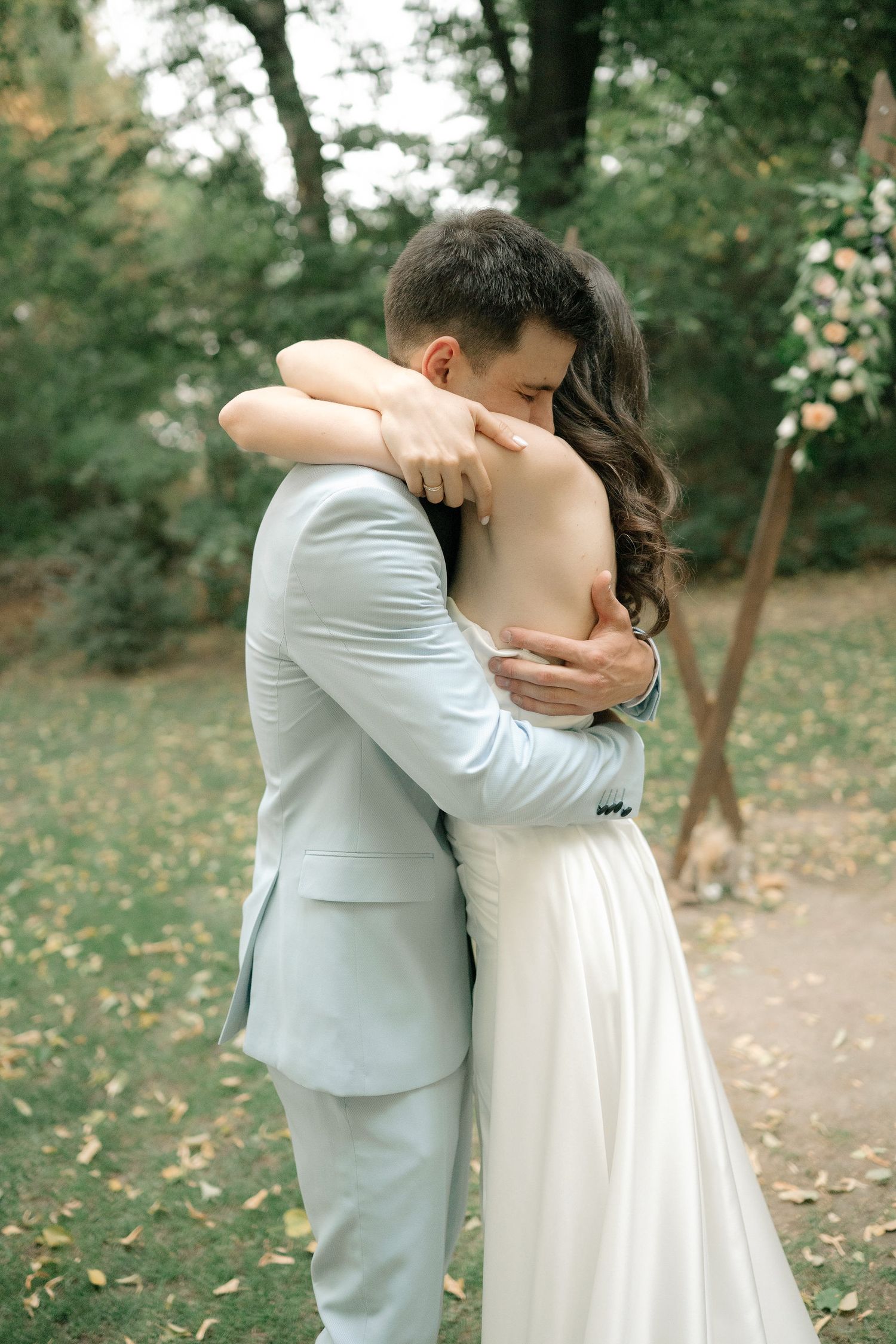 A tender embrace between a couple in wedding attire standing in a lush garden setting with autumn leaves scattered around.