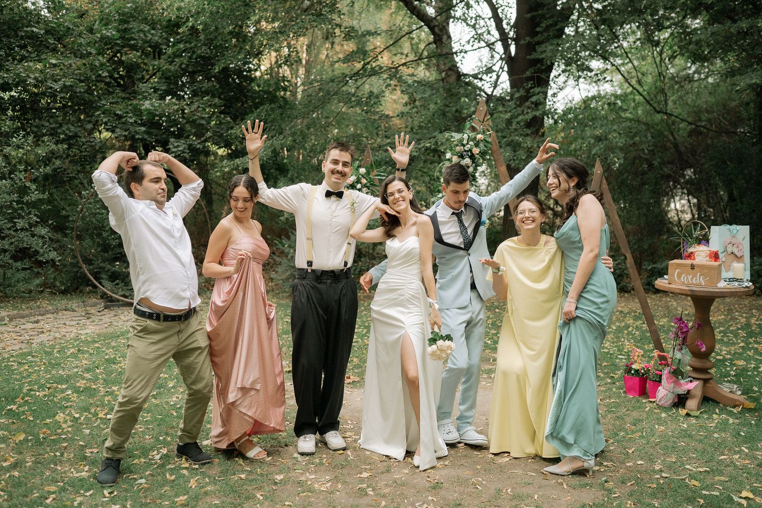 Wedding party in pastel dresses poses joyfully in a garden setting during summer celebration.