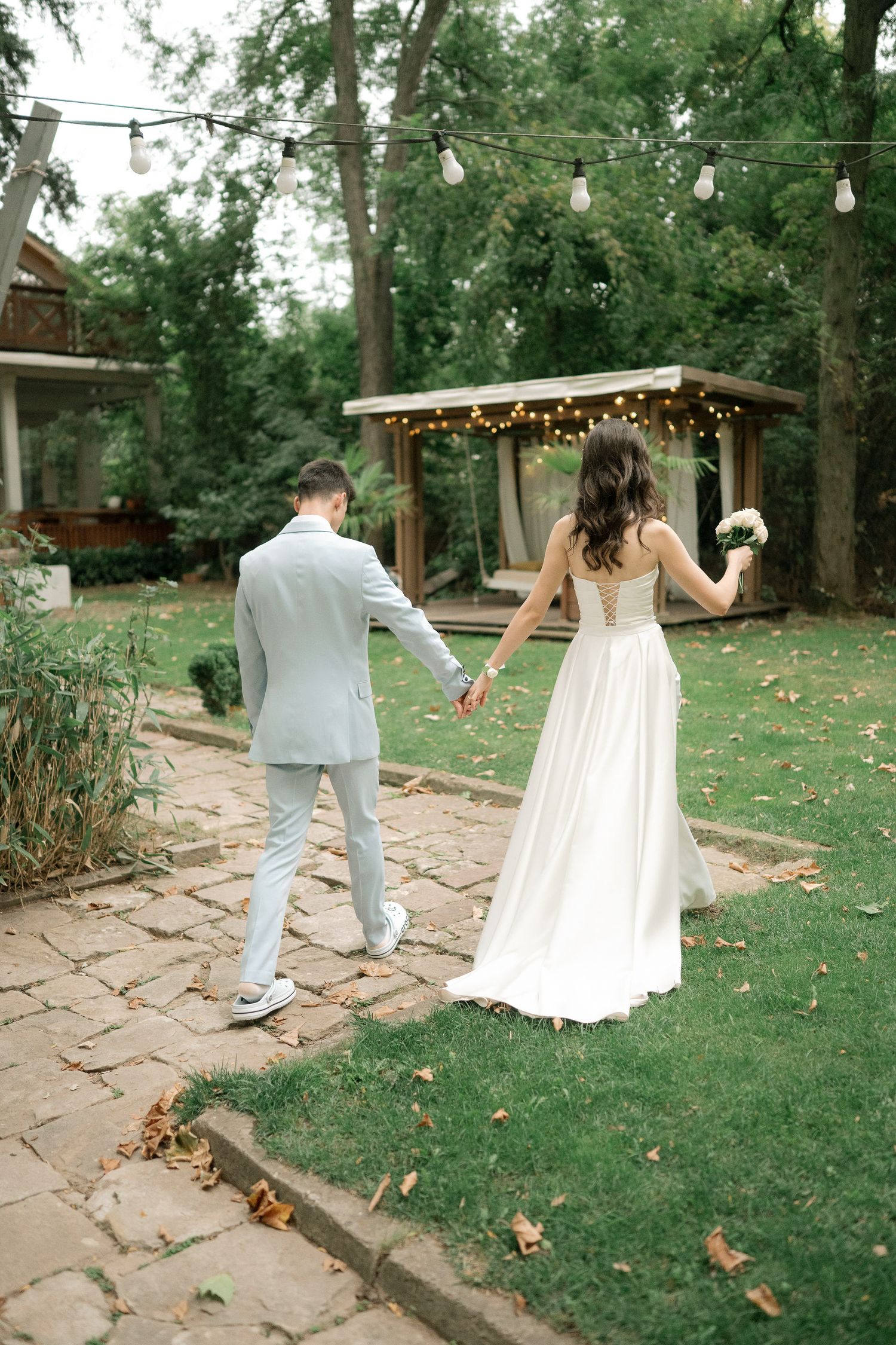 A couple in wedding attire walking through a garden pathway with string lights hanging overhead in a romantic setting.