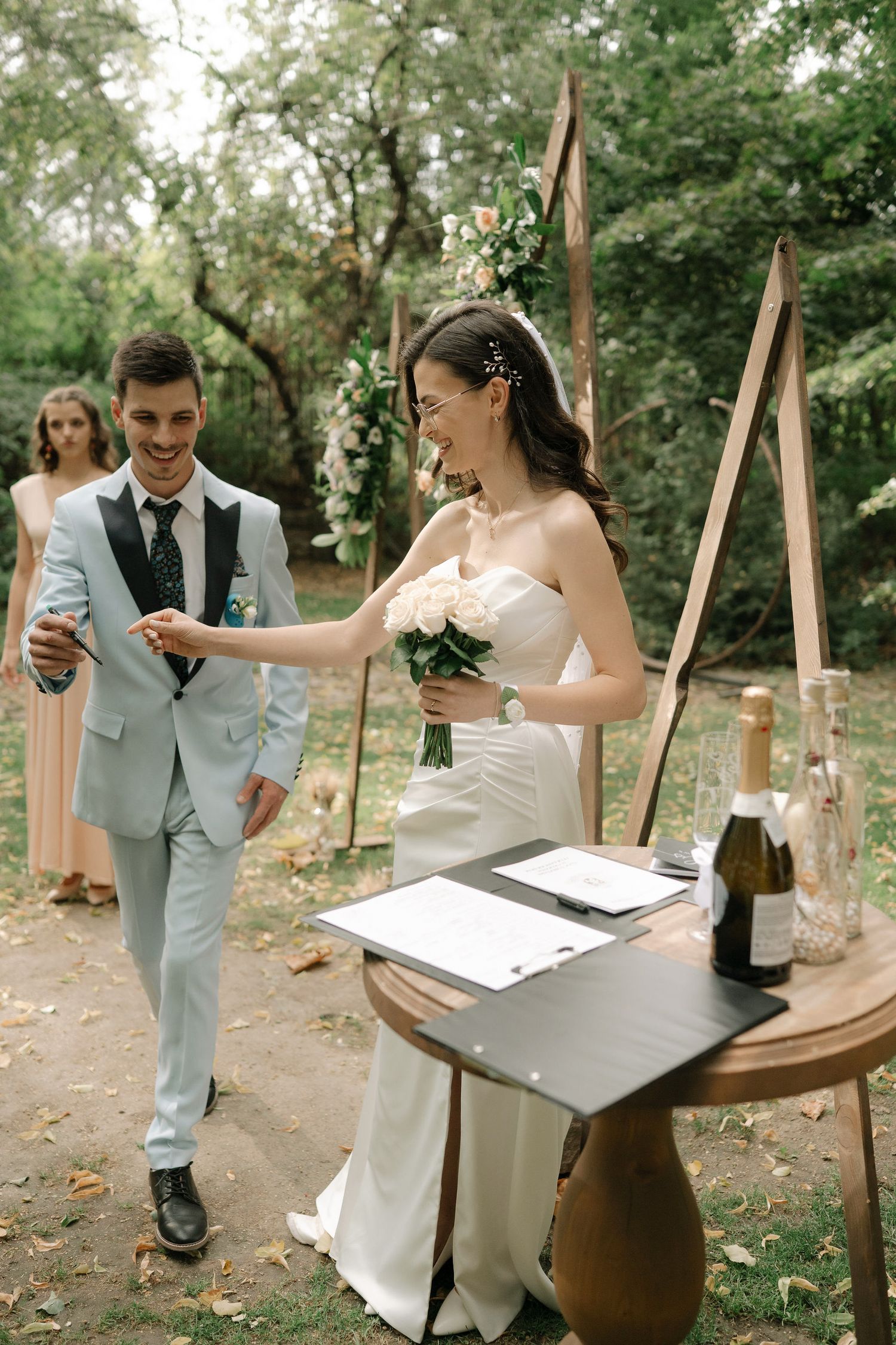 Couple signing marriage documents at outdoor wedding ceremony in wooded setting.