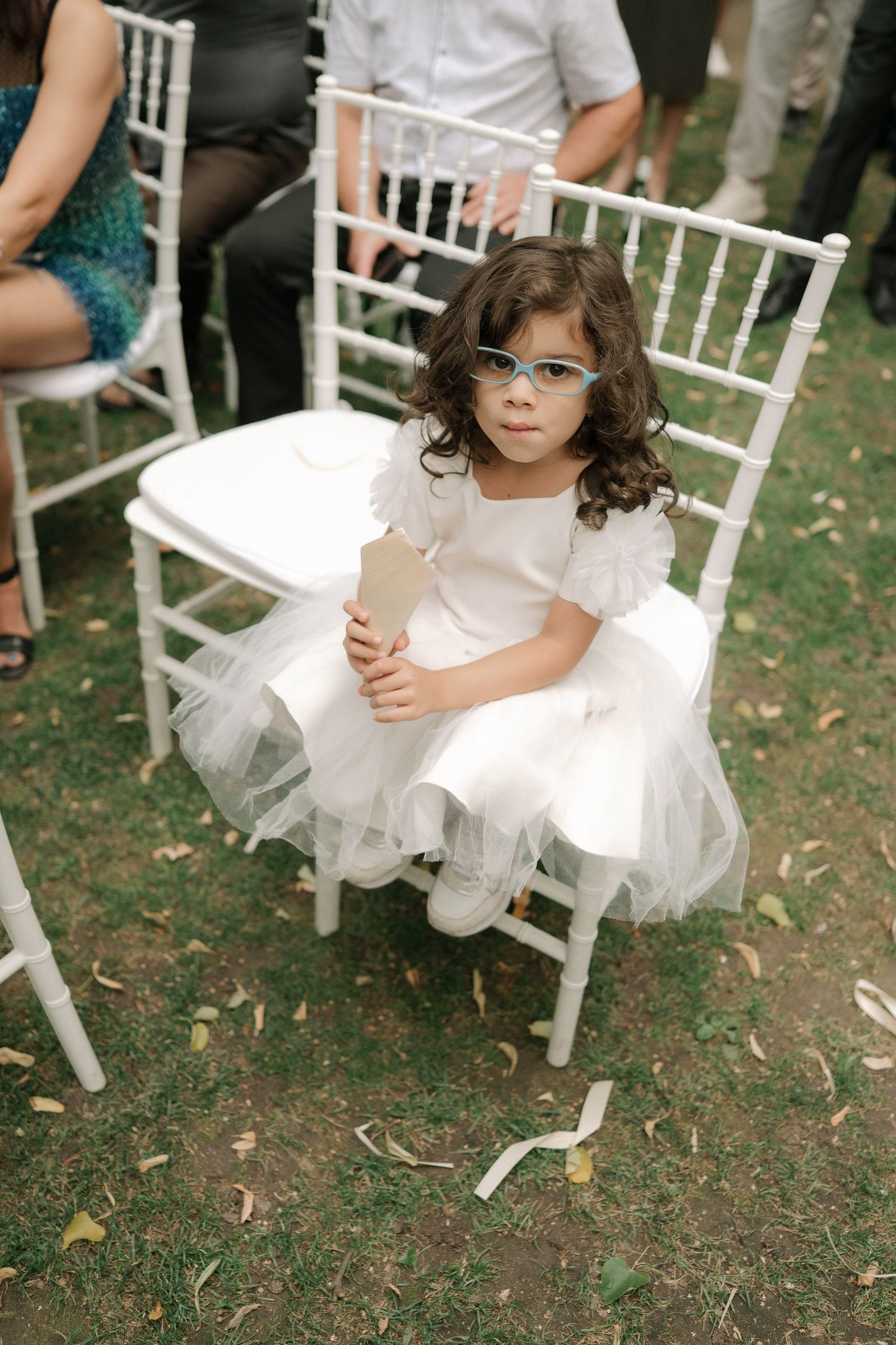 Young child in white dress sitting on white wooden chair at outdoor event with scattered leaves on ground.