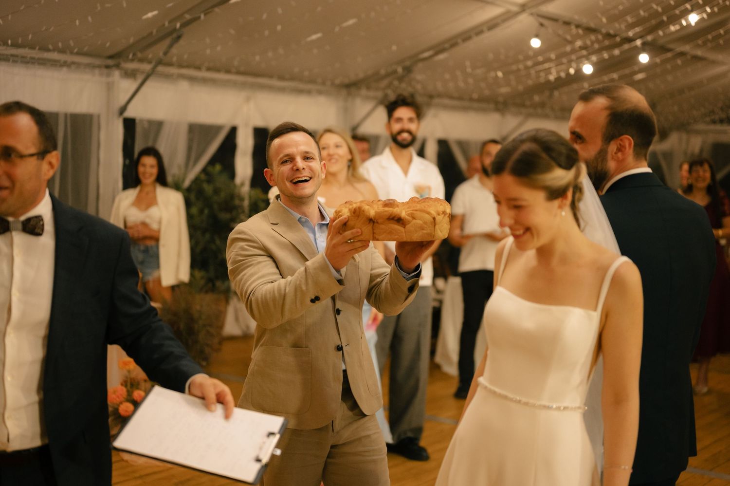 A couple shares a joyful moment during a wedding reception in a tent venue with warm lighting and guests in formal attire.