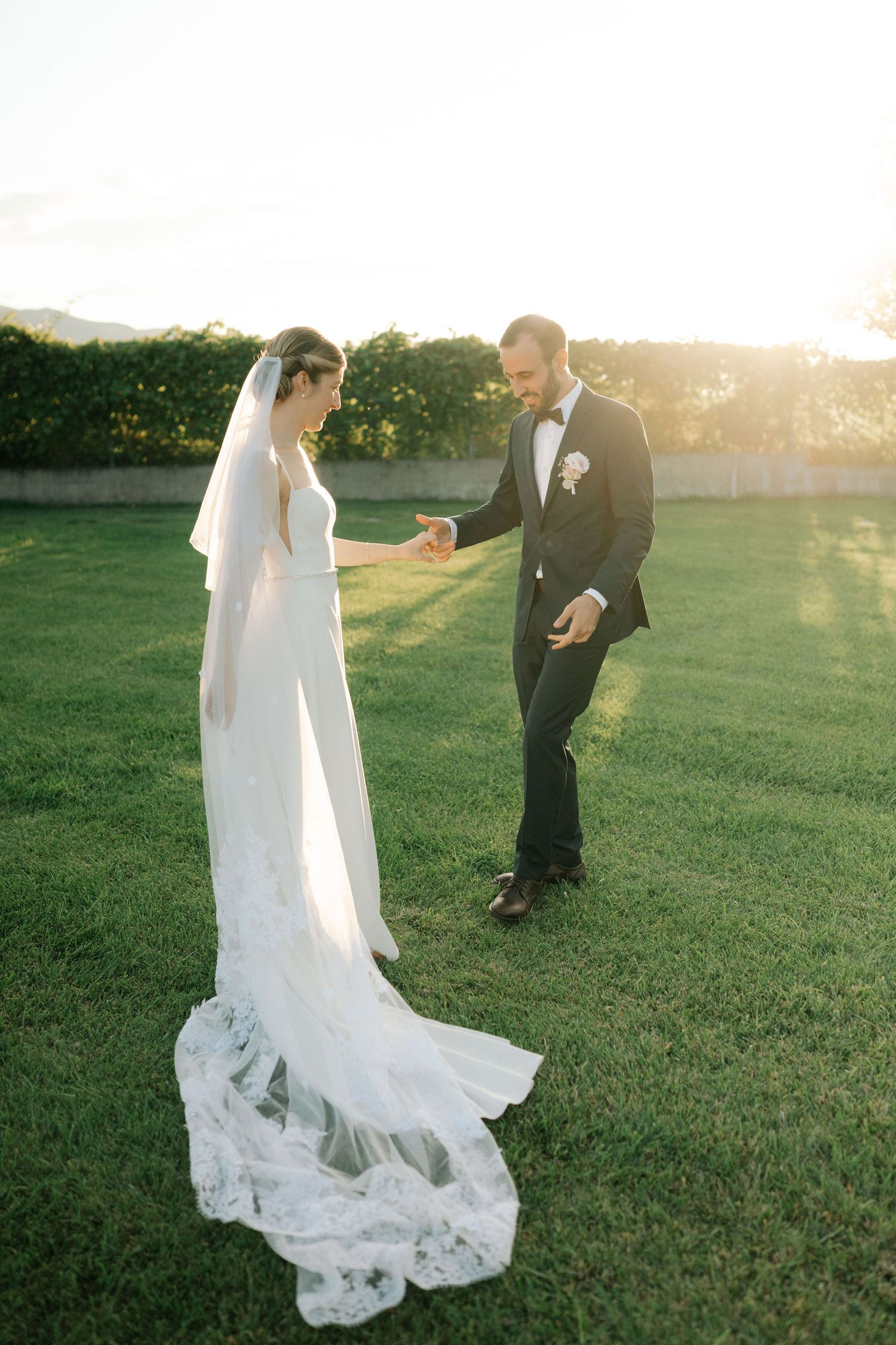 A wedding couple shares a romantic moment on a sunlit lawn with the bride's lace dress trailing behind.