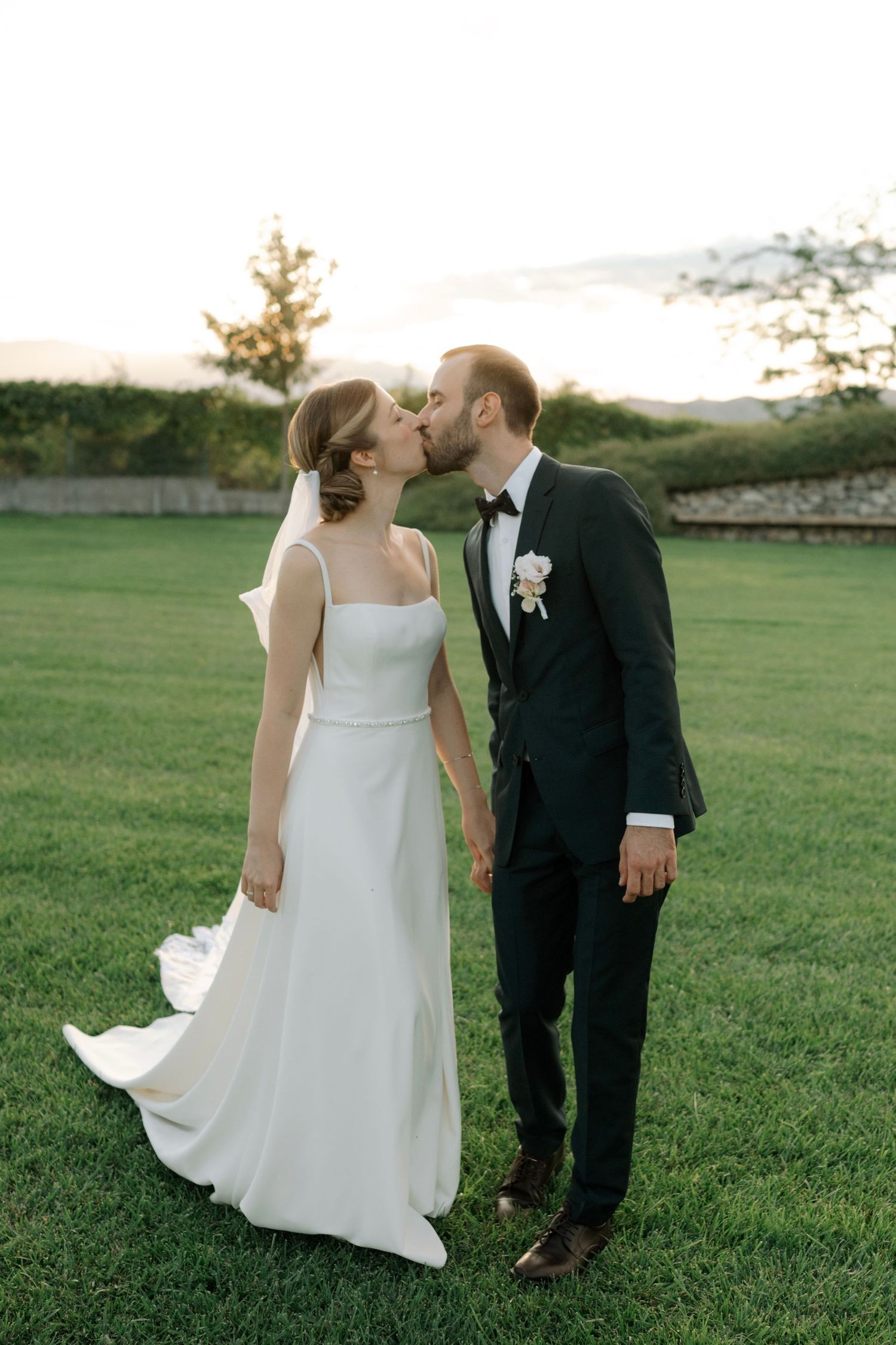 A romantic couple shares a kiss on a grassy lawn while wearing formal wedding attire at sunset.