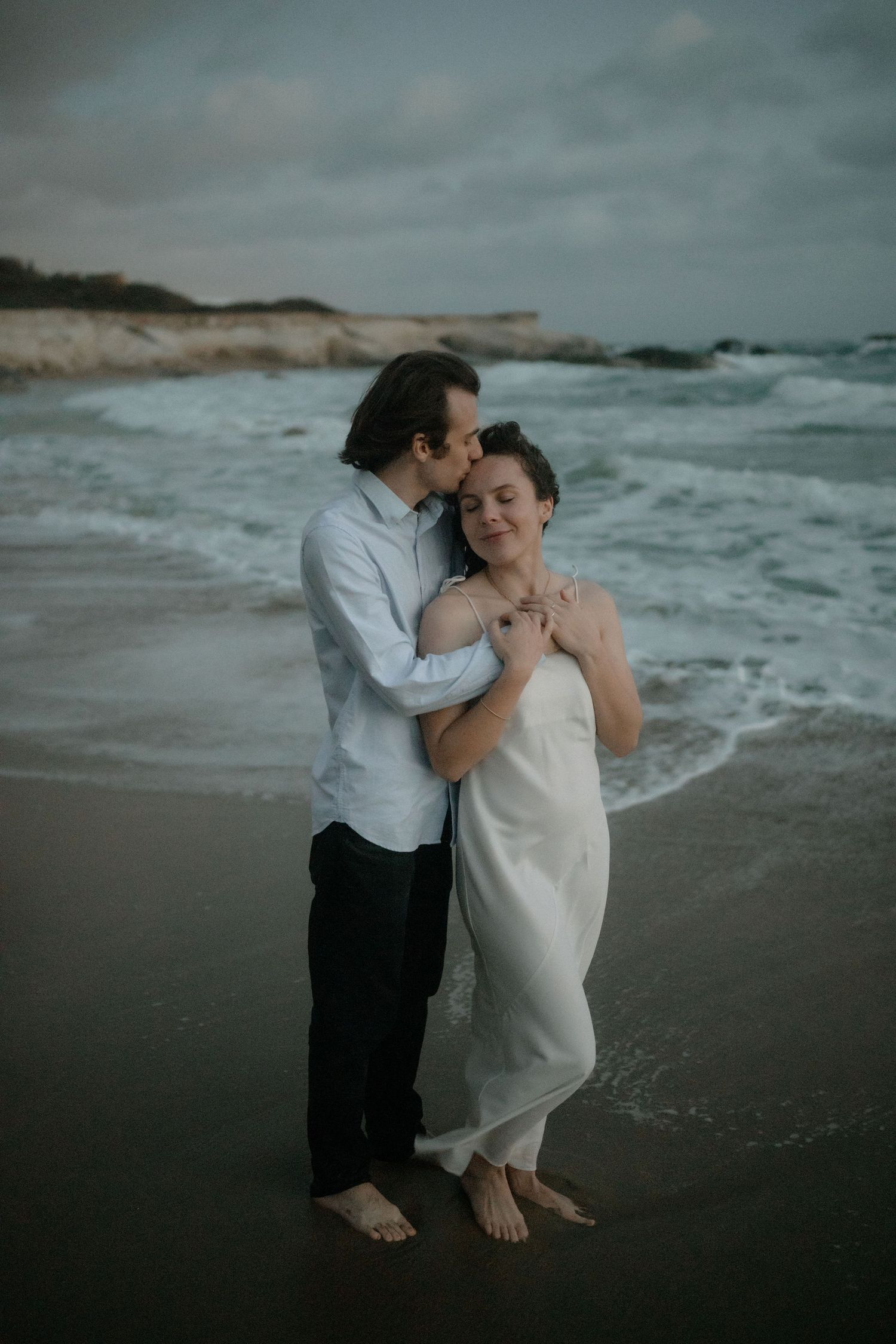 Couple embracing on a moody beach at dusk, man kissing woman's cheek, waves crashing behind them.