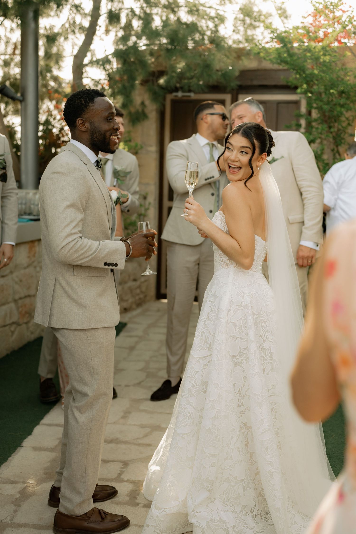 Bride in lace gown and groom in beige suit share a joyful moment at an outdoor wedding reception.