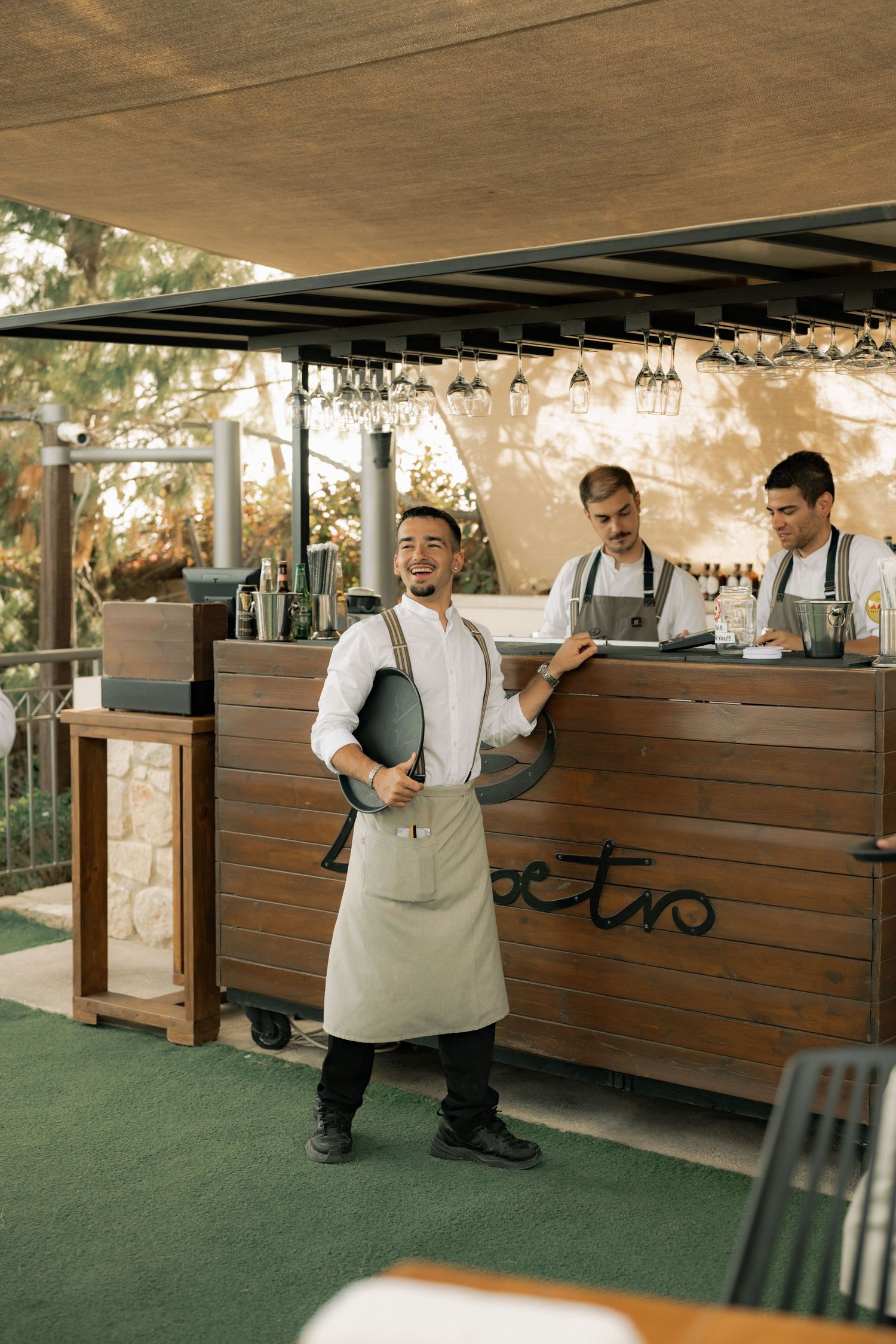 Smiling waiter in apron stands at outdoor restaurant bar with colleagues preparing drinks behind him.