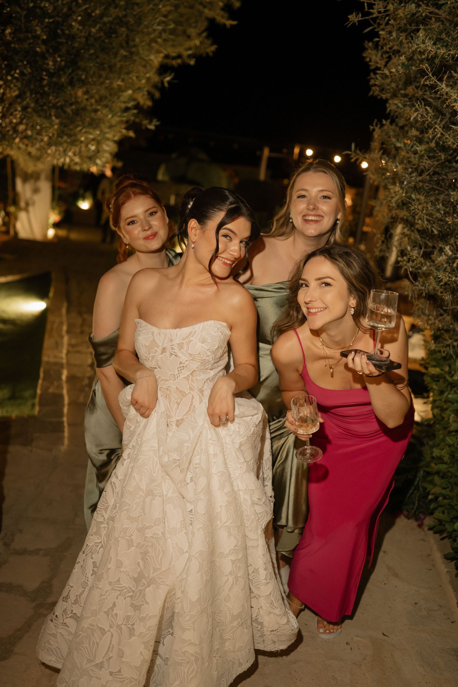Bride in white lace gown poses with three bridesmaids in sage and pink dresses at an outdoor evening wedding.