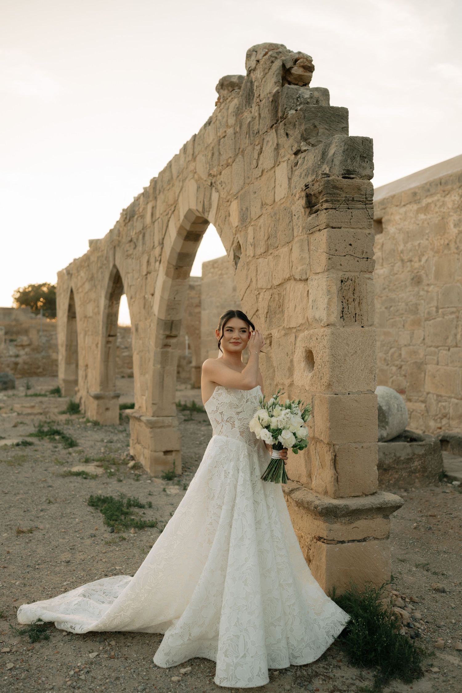 Bride in white lace gown holding bouquet poses before ancient stone arches at sunset.