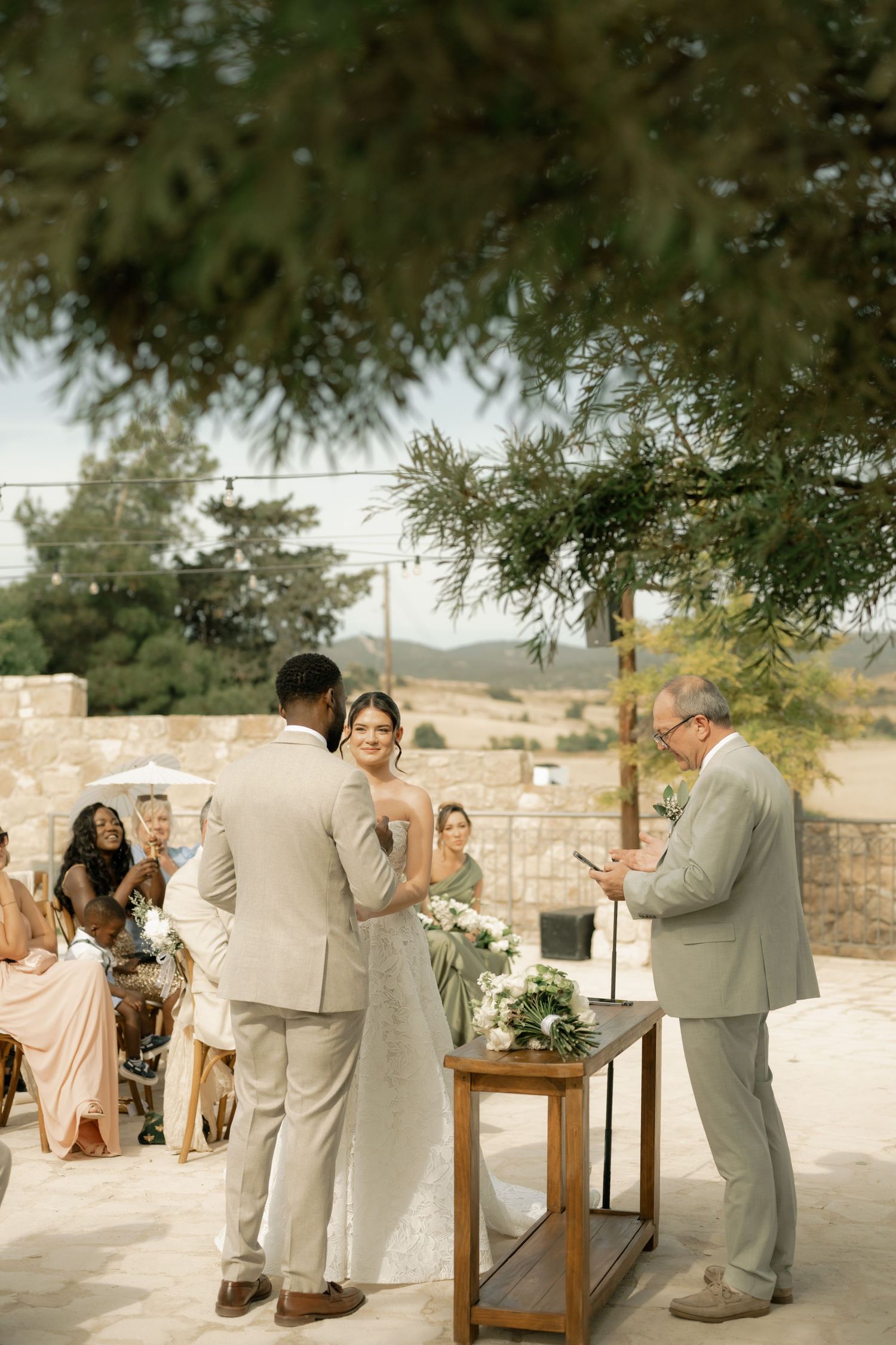 Outdoor wedding ceremony with bride and groom exchanging vows before an officiant, set against a scenic hillside backdrop.