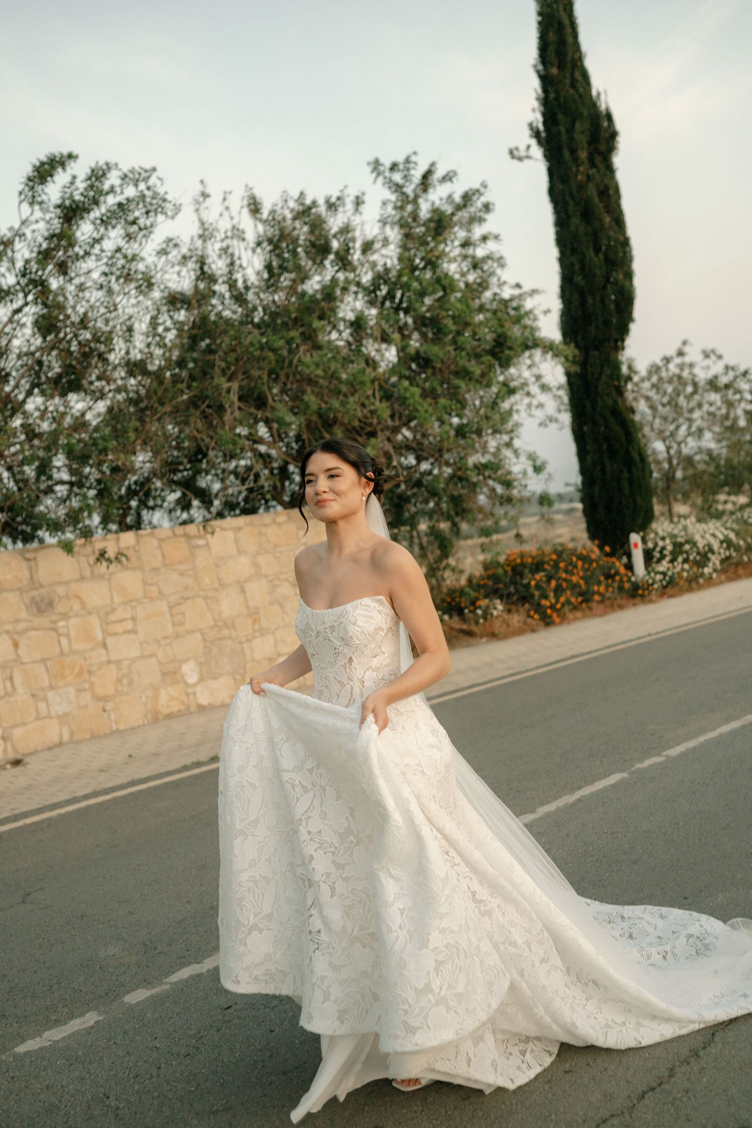 Bride in strapless lace wedding gown holding her train on a road, with cypress trees and stone wall backdrop.