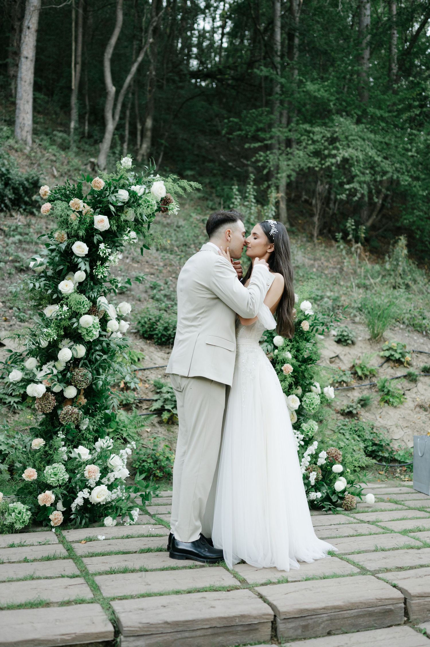 Newlyweds share an intimate moment against a backdrop of white roses and forest greenery.