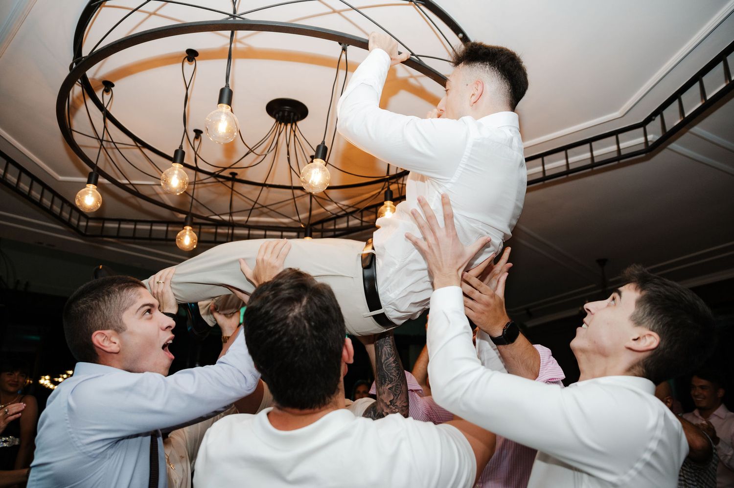 Wedding guests performing synchronized dance moves under ornate ceiling chandeliers.