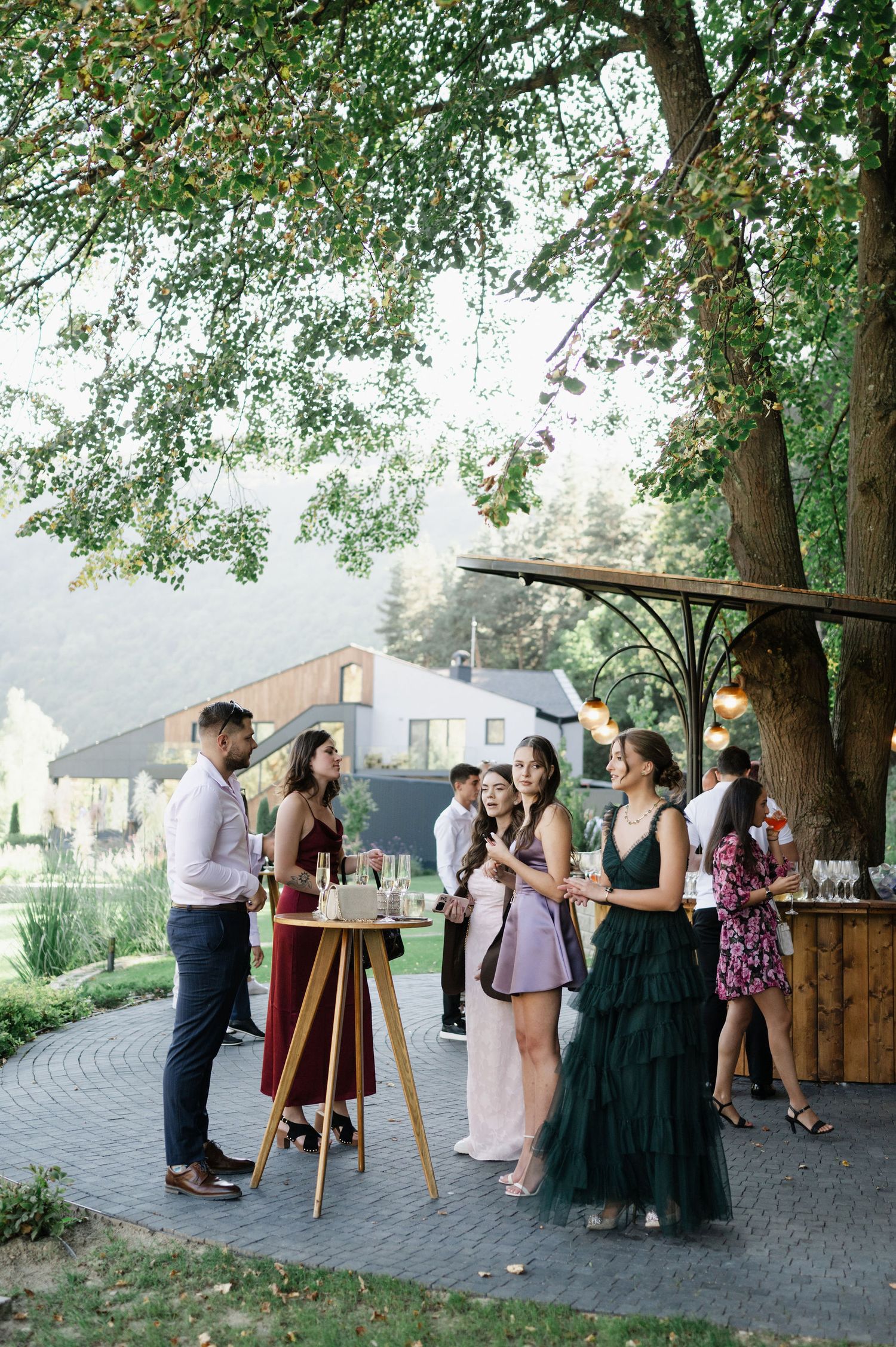 Wedding guests mingling at outdoor cocktail reception under tree canopy.