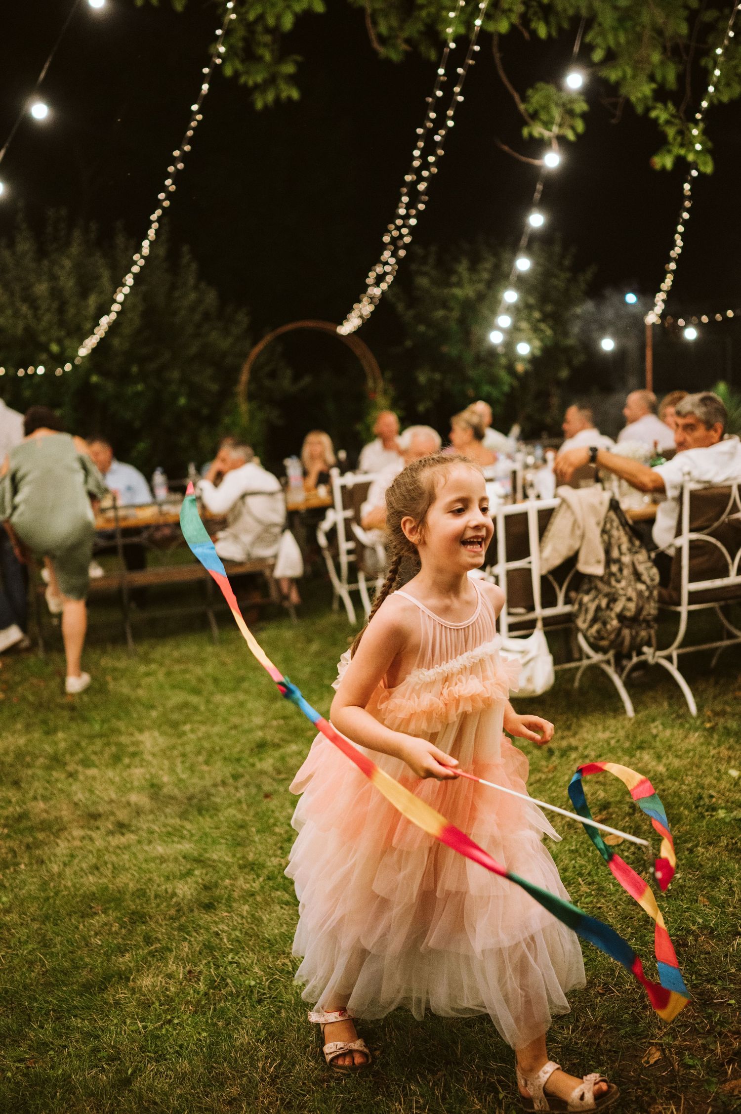 A young child in a peach tulle dress plays with ribbons under string lights at an outdoor party.