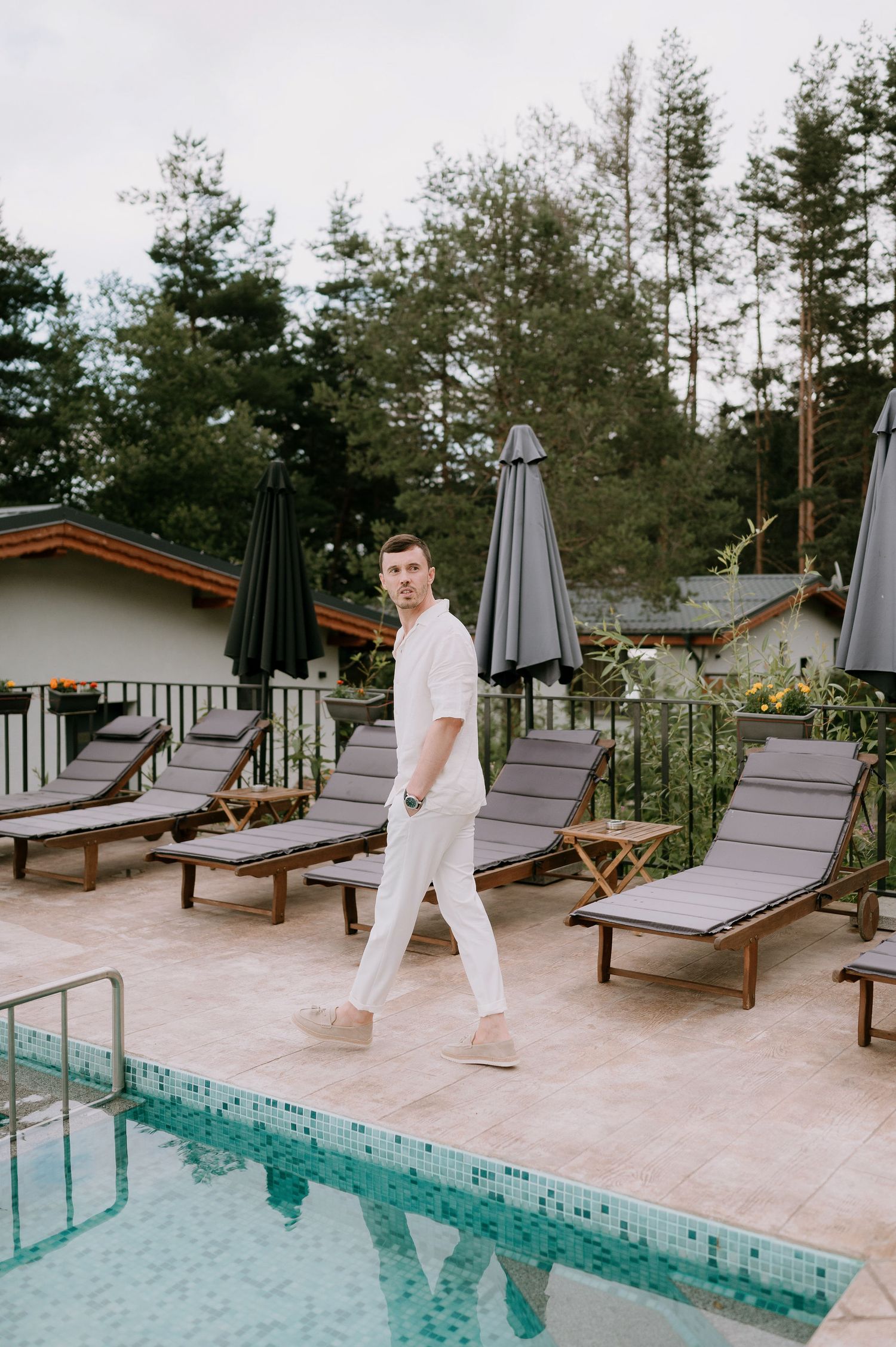 Person in white walking along a pool deck with lounge chairs and pine trees in background.