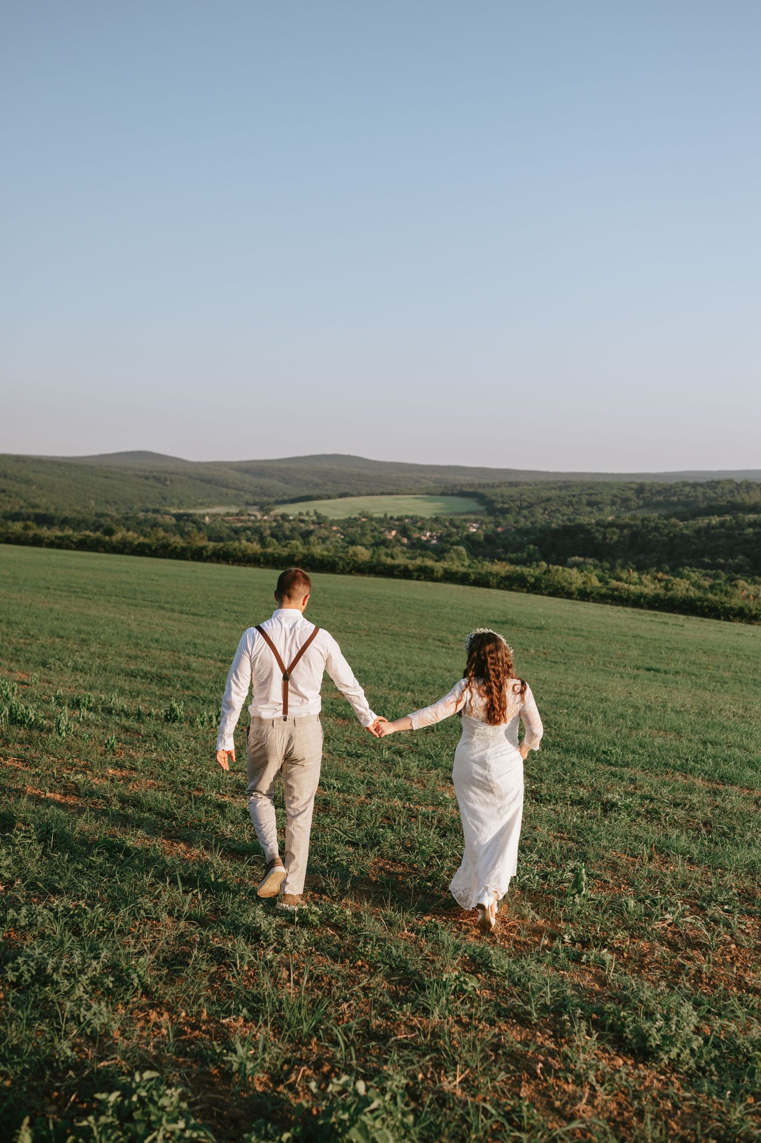 Newlyweds walk hand in hand through a vast green hillside landscape.