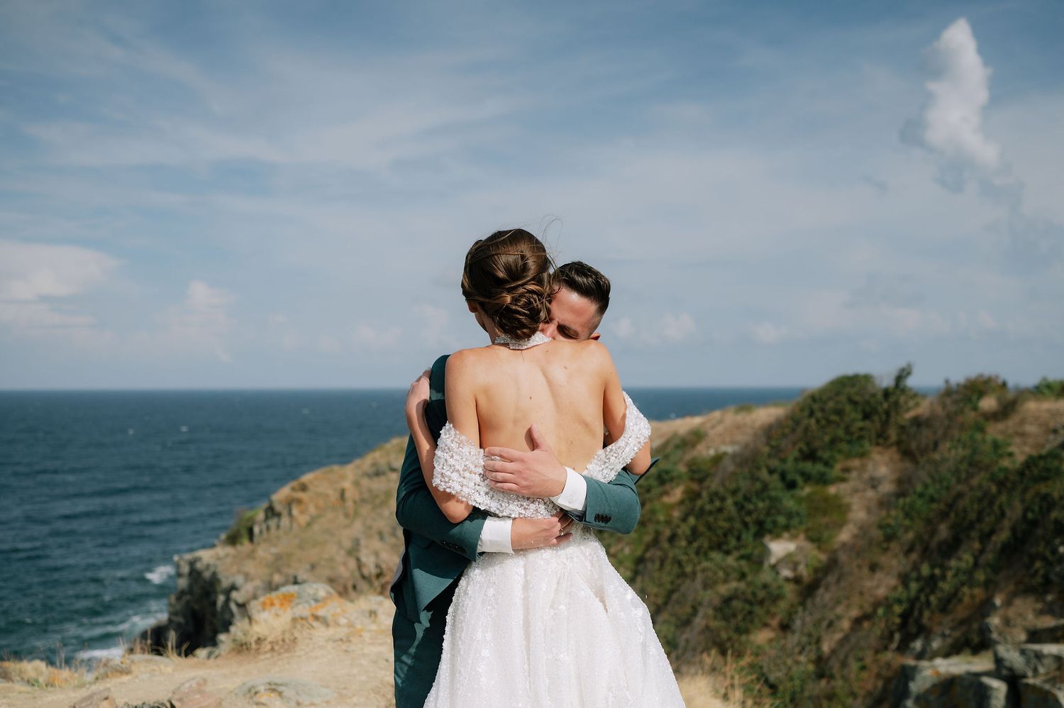A romantic wedding photoshoot on a dramatic coastal cliff overlooking the ocean on a sunny day.