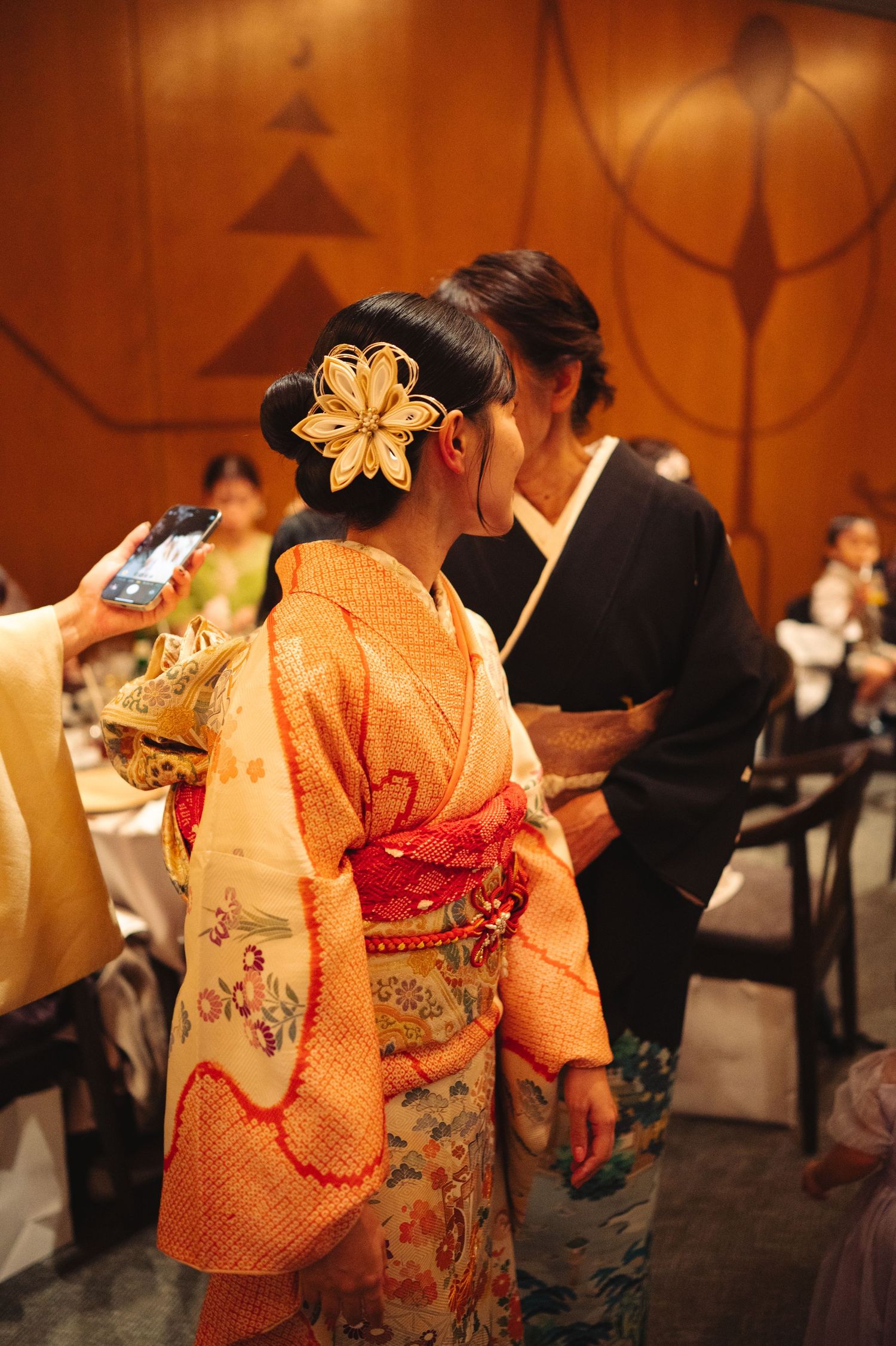 Women in vibrant orange and red kimonos with floral patterns dance at a formal event.