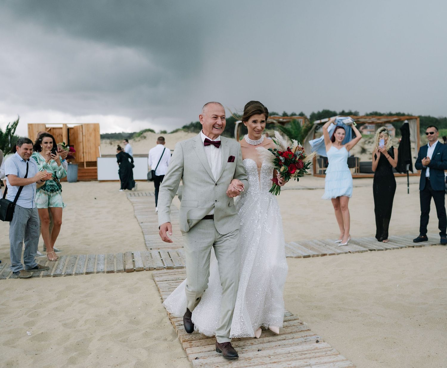Wedding party and guests line up along a wooden beach walkway during an overcast day.