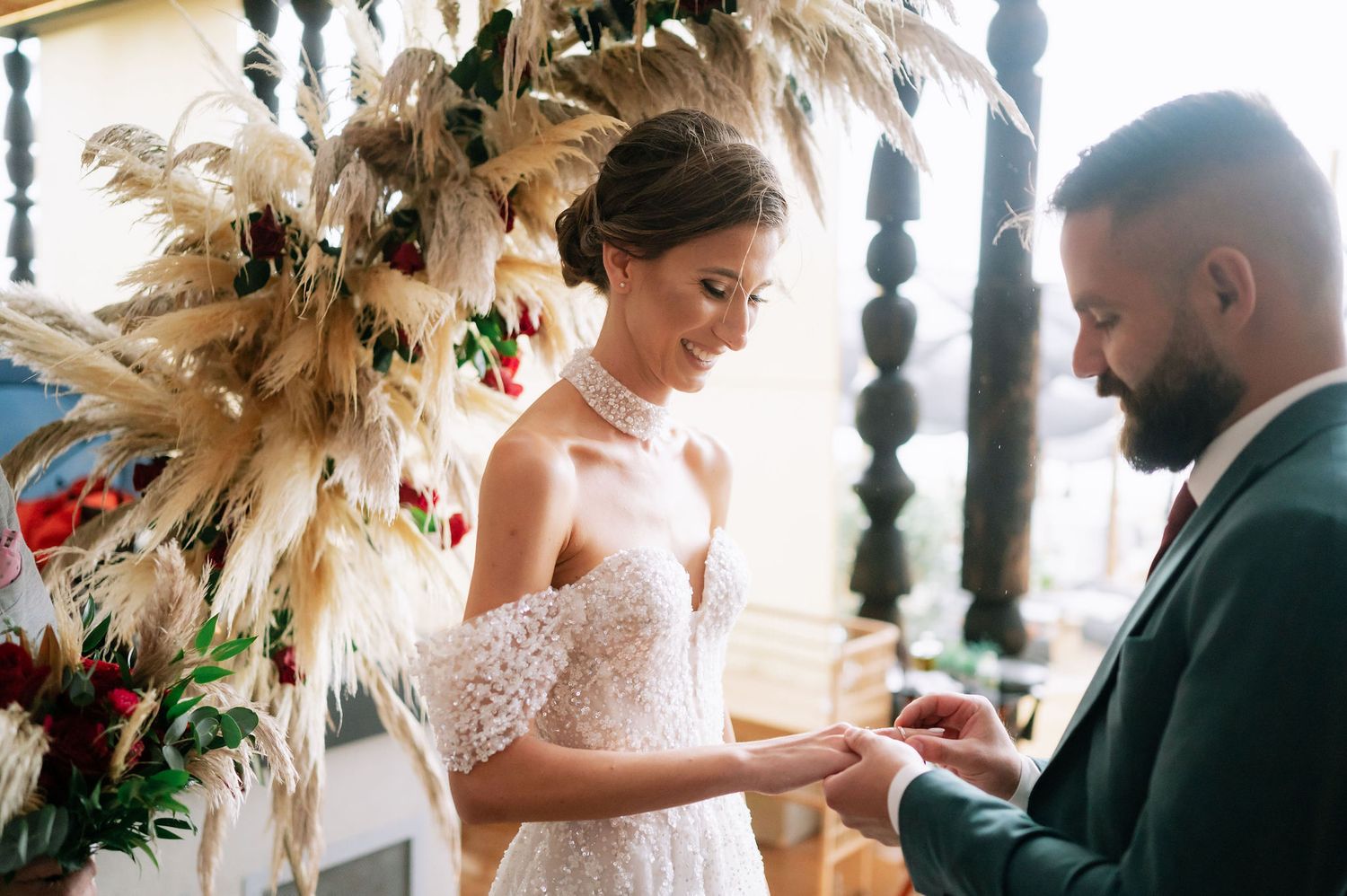 A romantic wedding ceremony moment with a white lace dress and dried pampas grass decor in a sunlit venue.