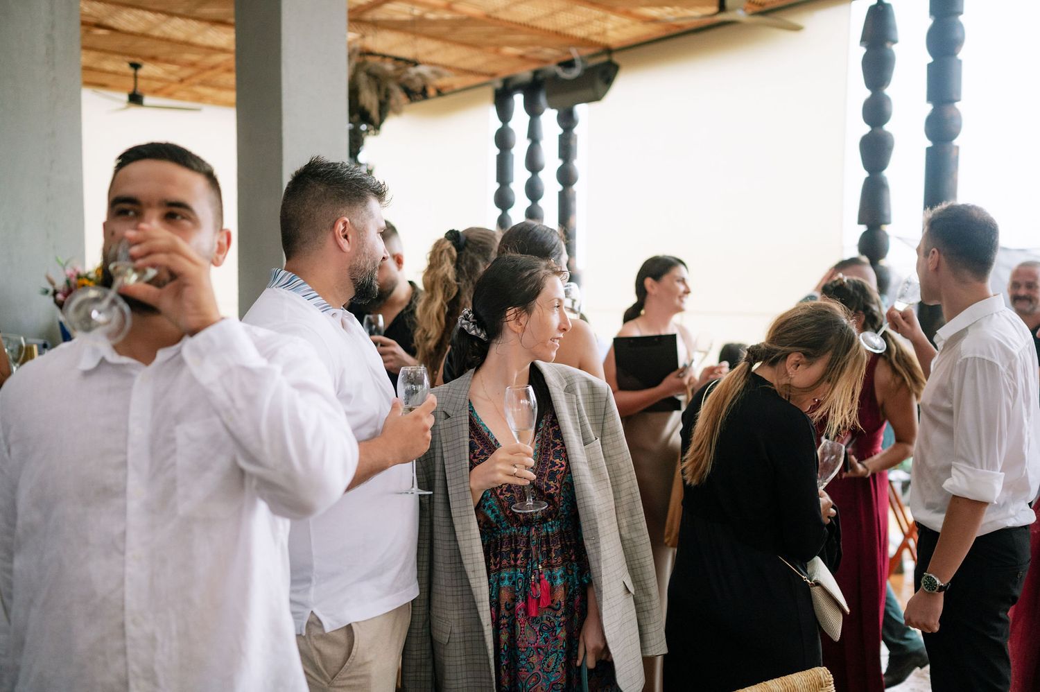 Wedding guests mingle and socialize at an indoor reception venue with rustic wooden ceiling beams.