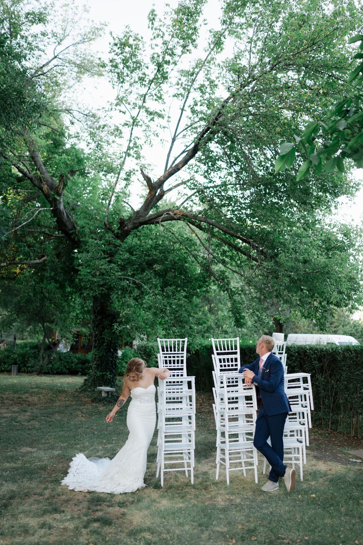 A couple interacts playfully among white ceremony chairs in a garden setting.