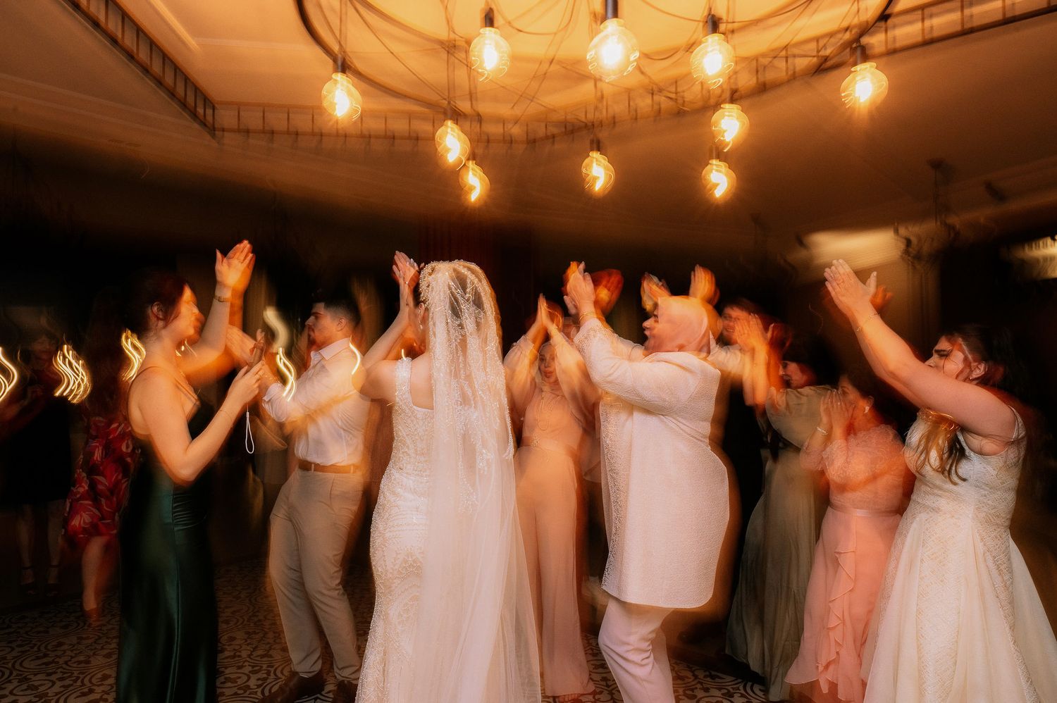 Wedding guests dance and celebrate on the dance floor under warm glowing lights at an evening reception.