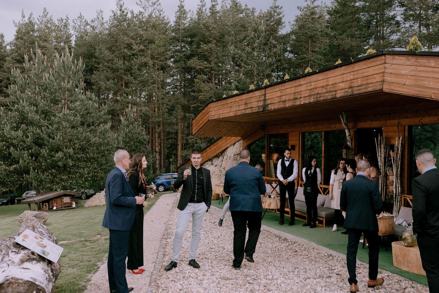 Wedding guests gathered around a rustic wooden pavilion in a mountain setting.