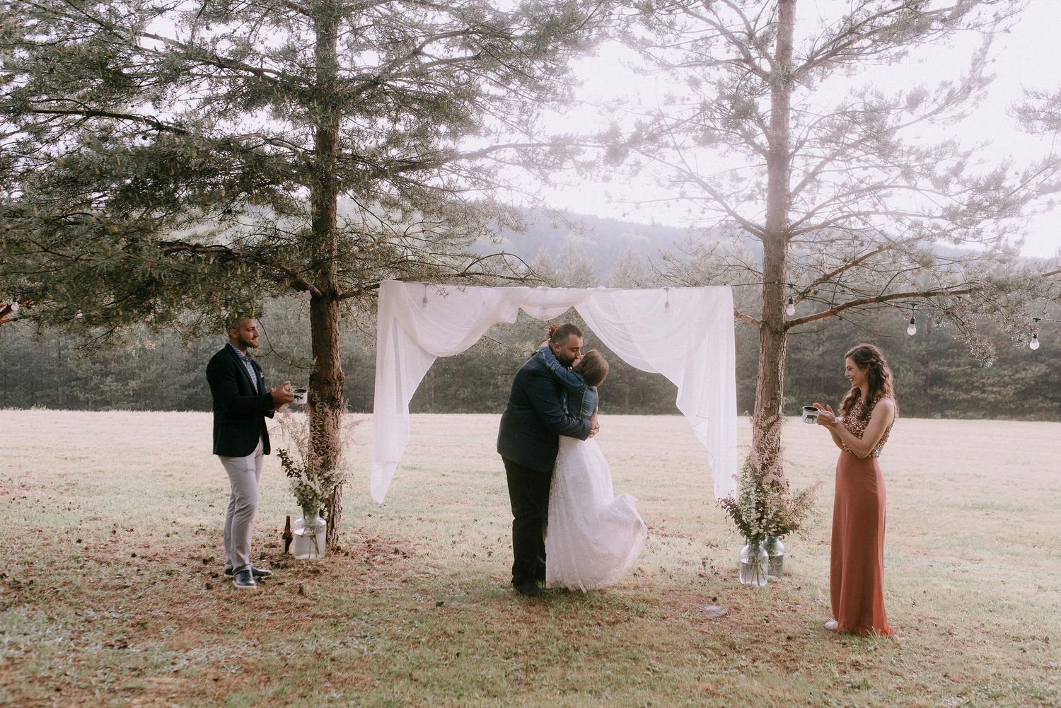 Wedding ceremony site with white fabric arch set against pine trees and rolling hills.