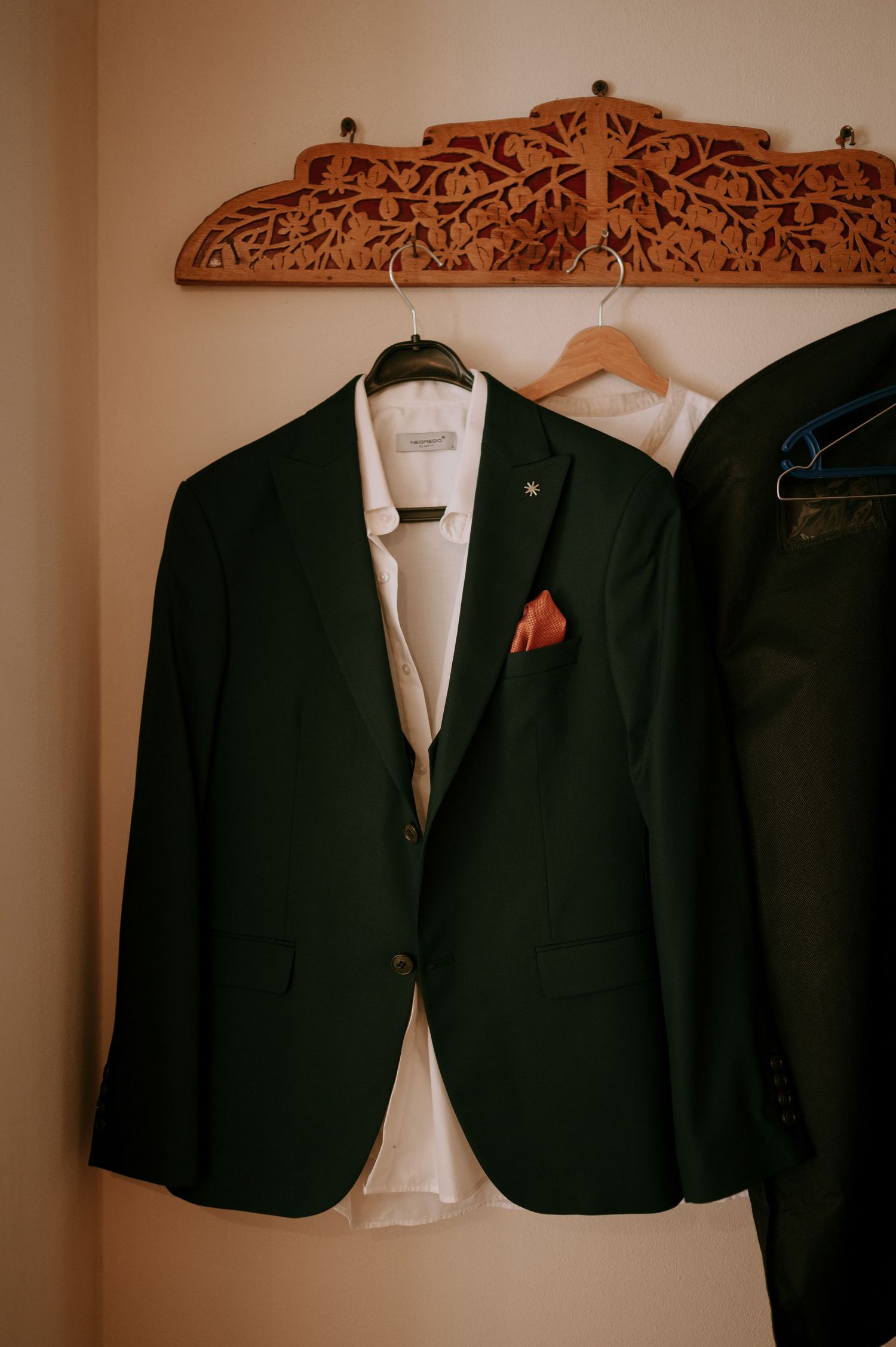 Dark green suit jacket with red pocket square hanging on wooden carved hanger.