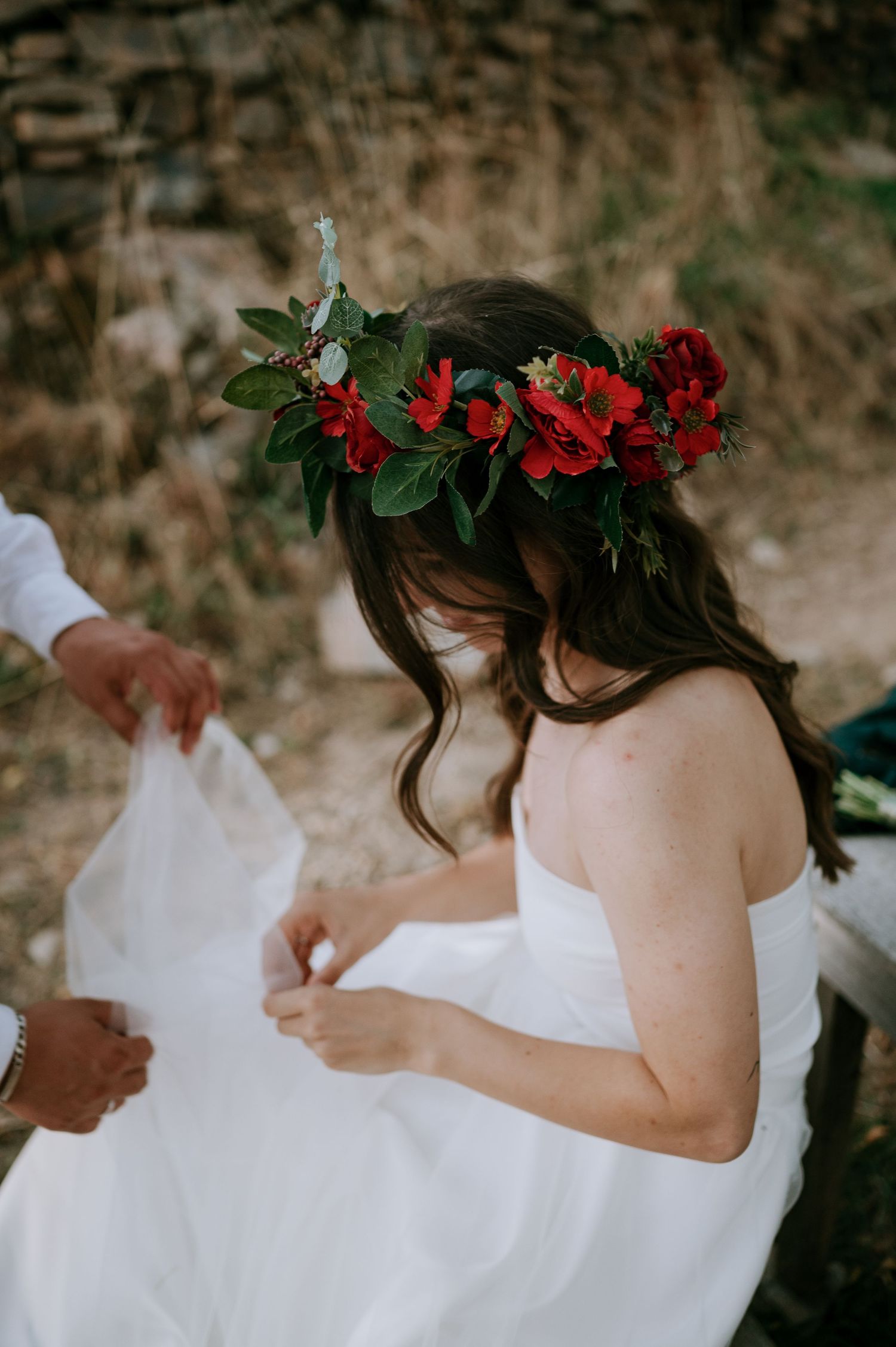A bride in a flowing white dress adorned with a red flower crown poses for artistic photos.