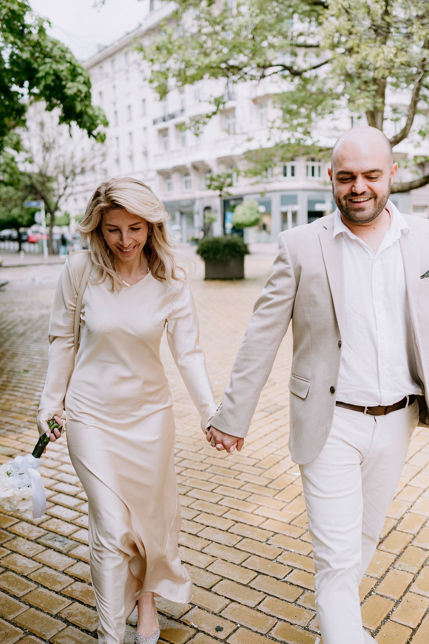 Happy couple in matching cream outfits walking hand in hand on brick pavement.