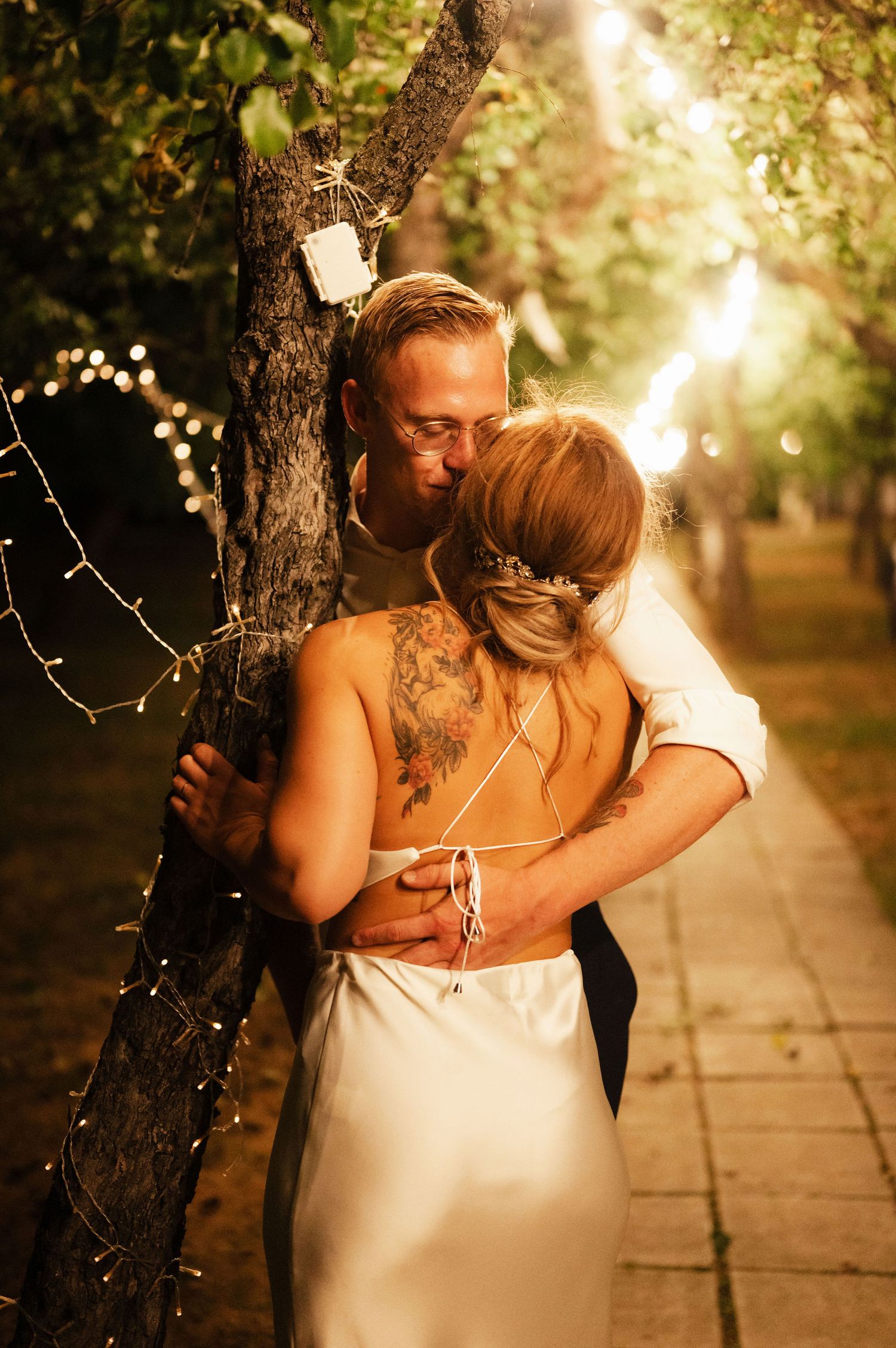A romantic couple embraces under glowing string lights in an outdoor garden setting at sunset.