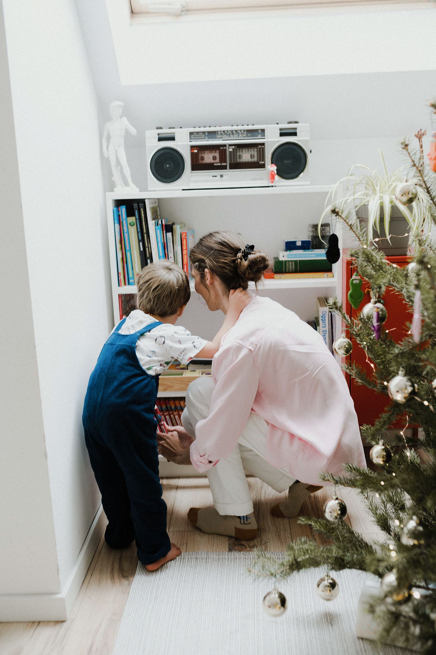 Two people looking at something together near a bookshelf with a retro boombox and Christmas tree in view.