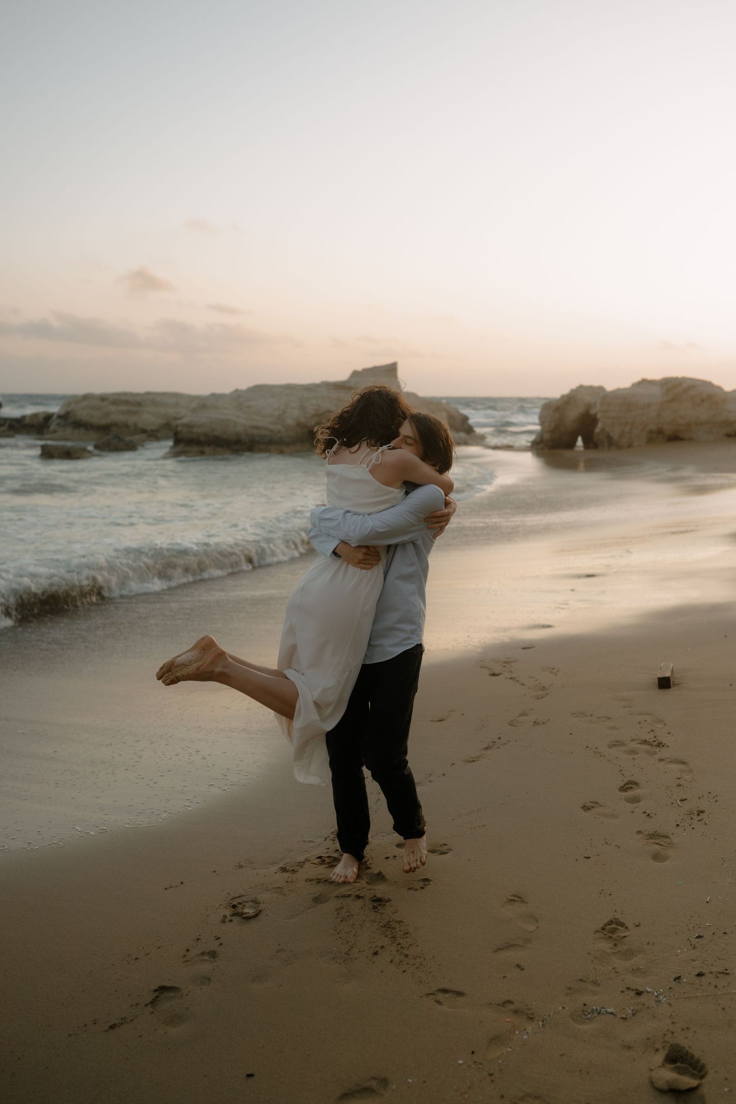 Couple embracing on a sandy beach at sunset with ocean waves and rocky cliffs in the background.