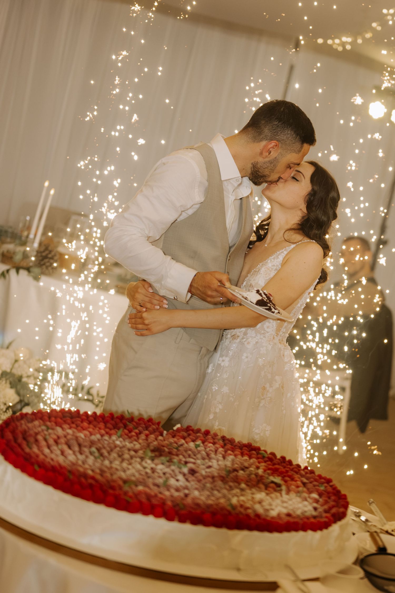 Newlyweds sharing a kiss while cutting their heart-shaped strawberry wedding cake surrounded by sparkling fireworks.