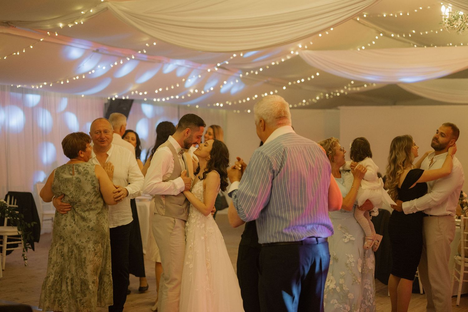 Couples dancing at a wedding reception under fairy lights and white draped ceiling decorations.