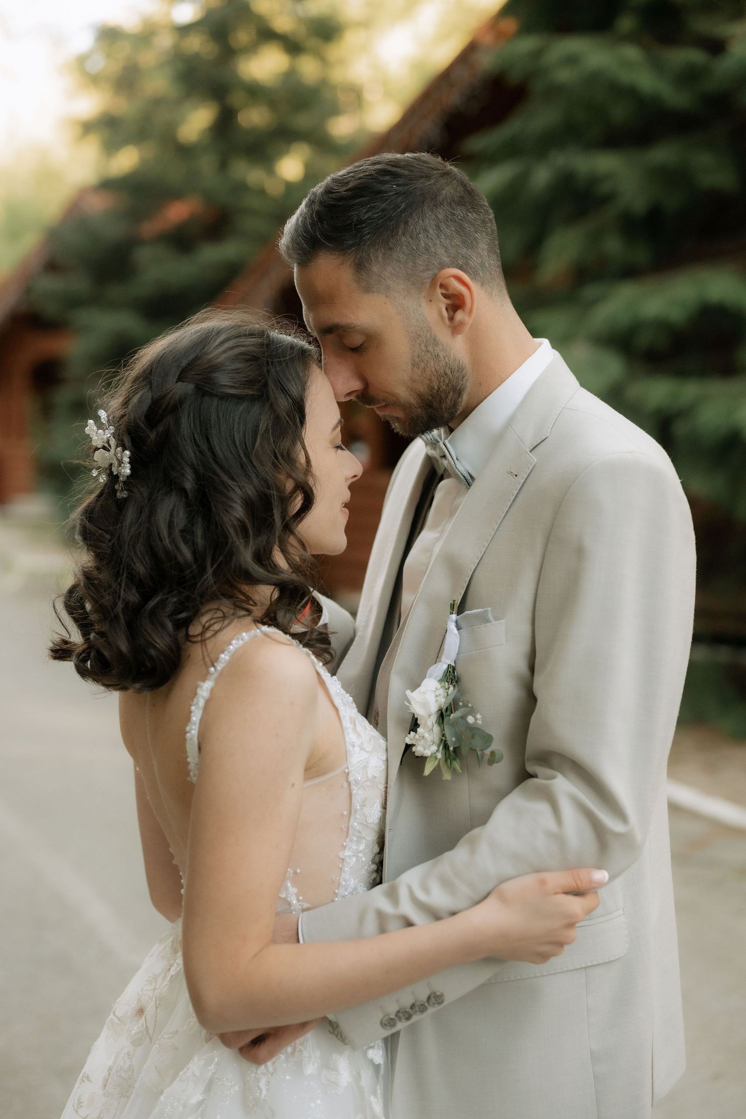 A bride and groom share a romantic moment, nearly kissing outdoors, dressed in wedding attire with greenery in the background.