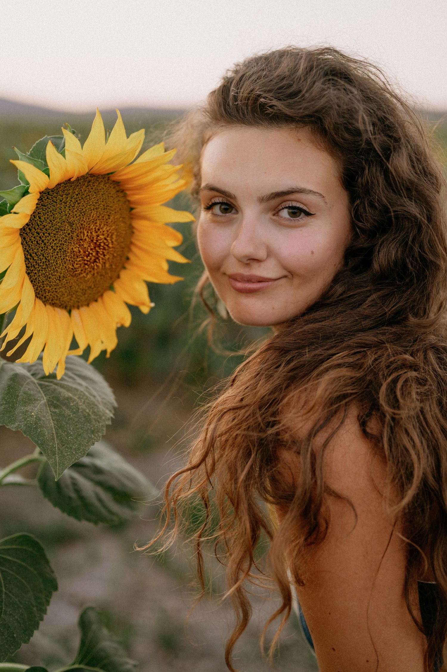 Young woman with curly brown hair smiling beside a bright yellow sunflower in an outdoor field at dusk.
