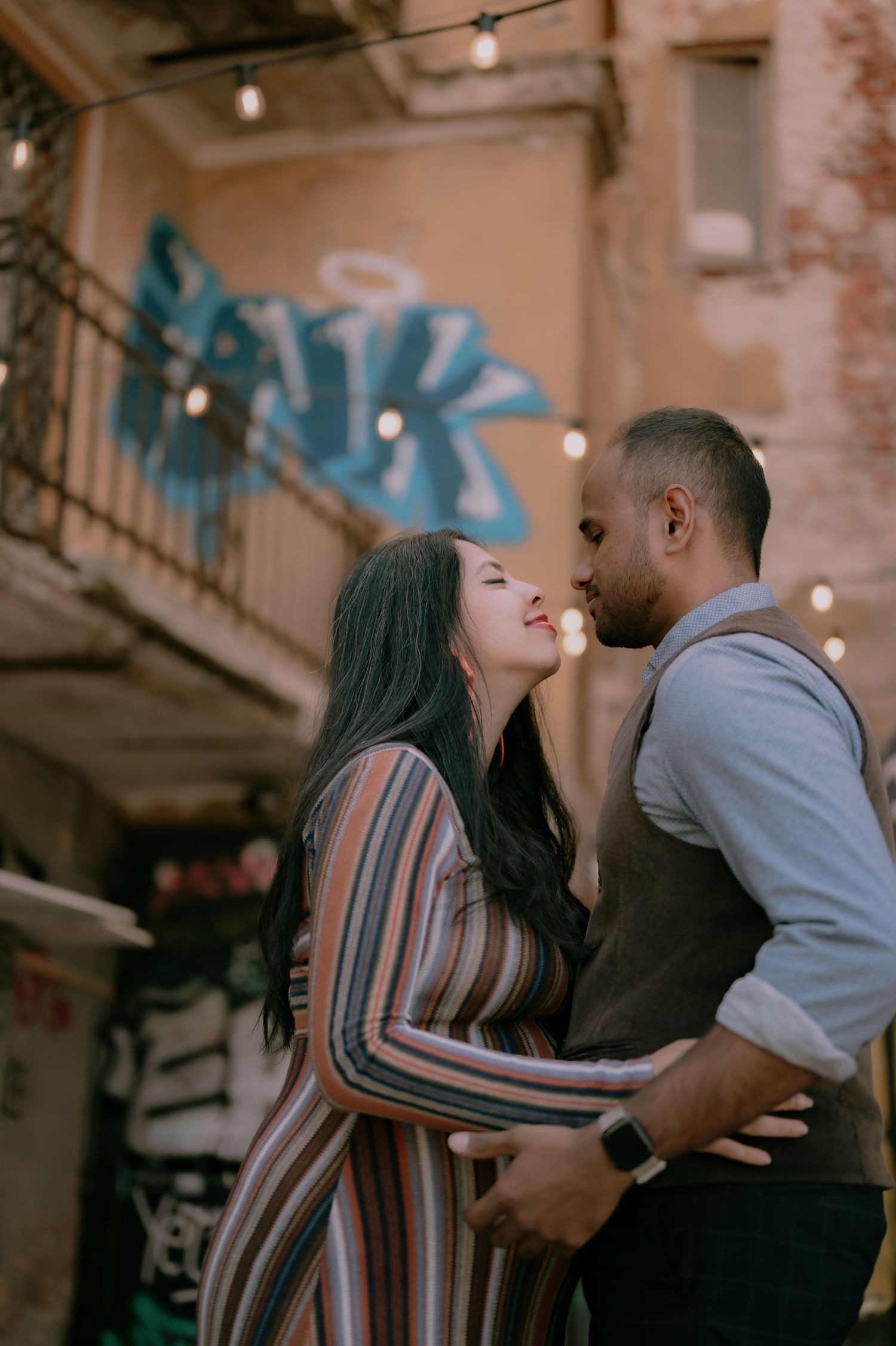 A couple shares a romantic moment in an urban alley, the woman in a striped dress gazing up at her partner amid string lights.