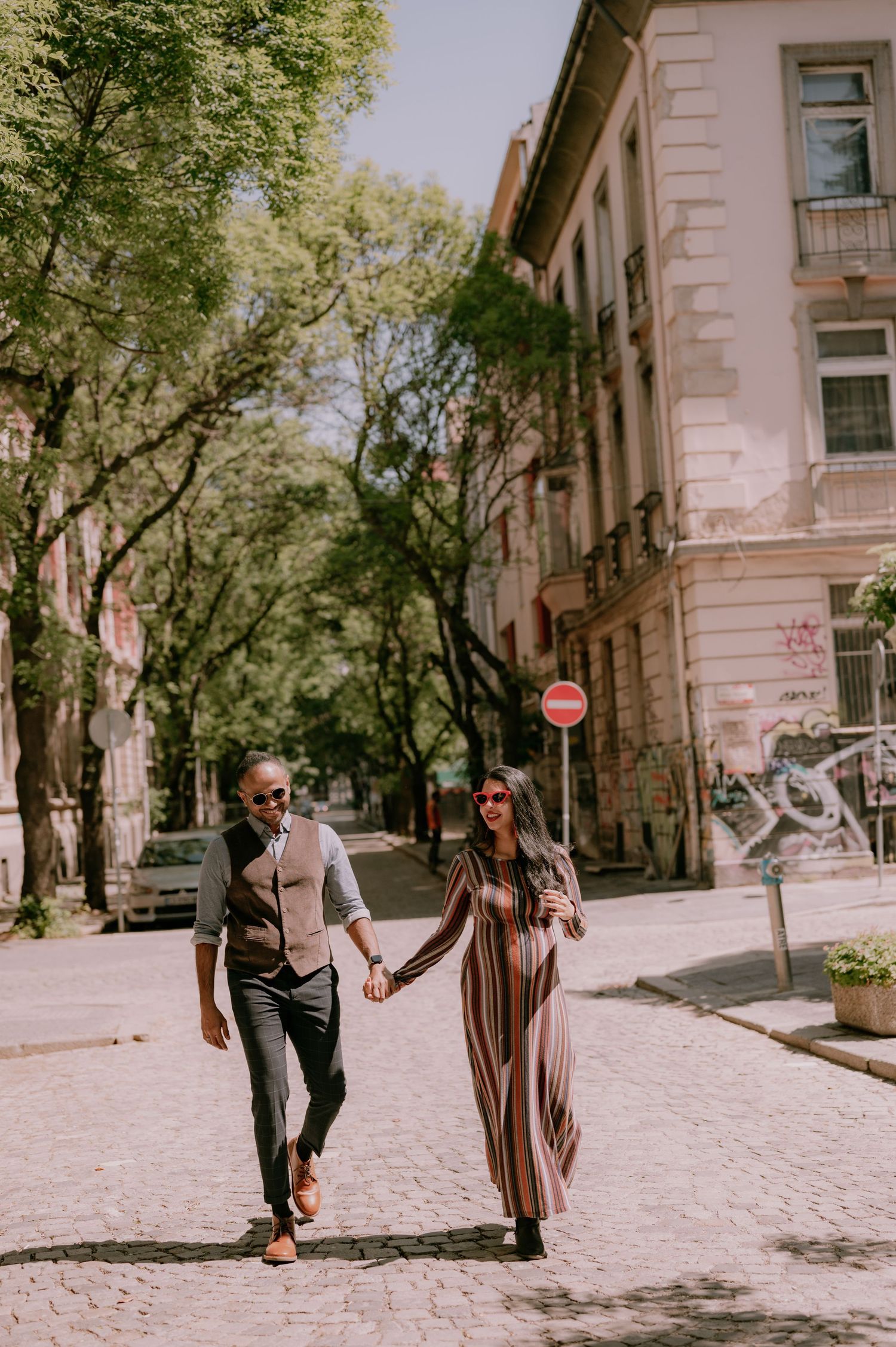 Couple holding hands walking on cobblestone street lined with trees and historic buildings on a sunny day.