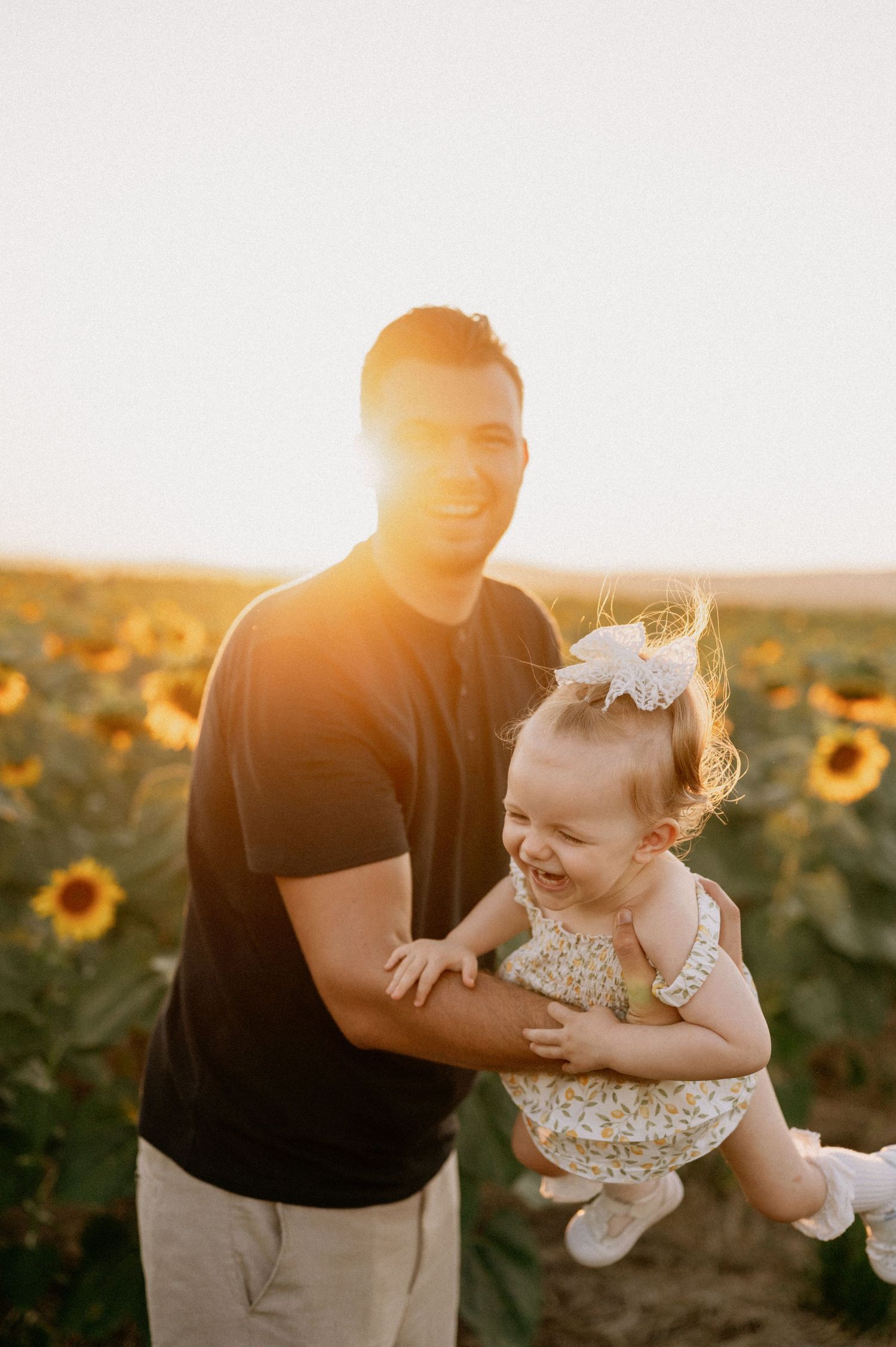 A smiling man holds a laughing baby girl in a sunflower field at golden hour sunset.